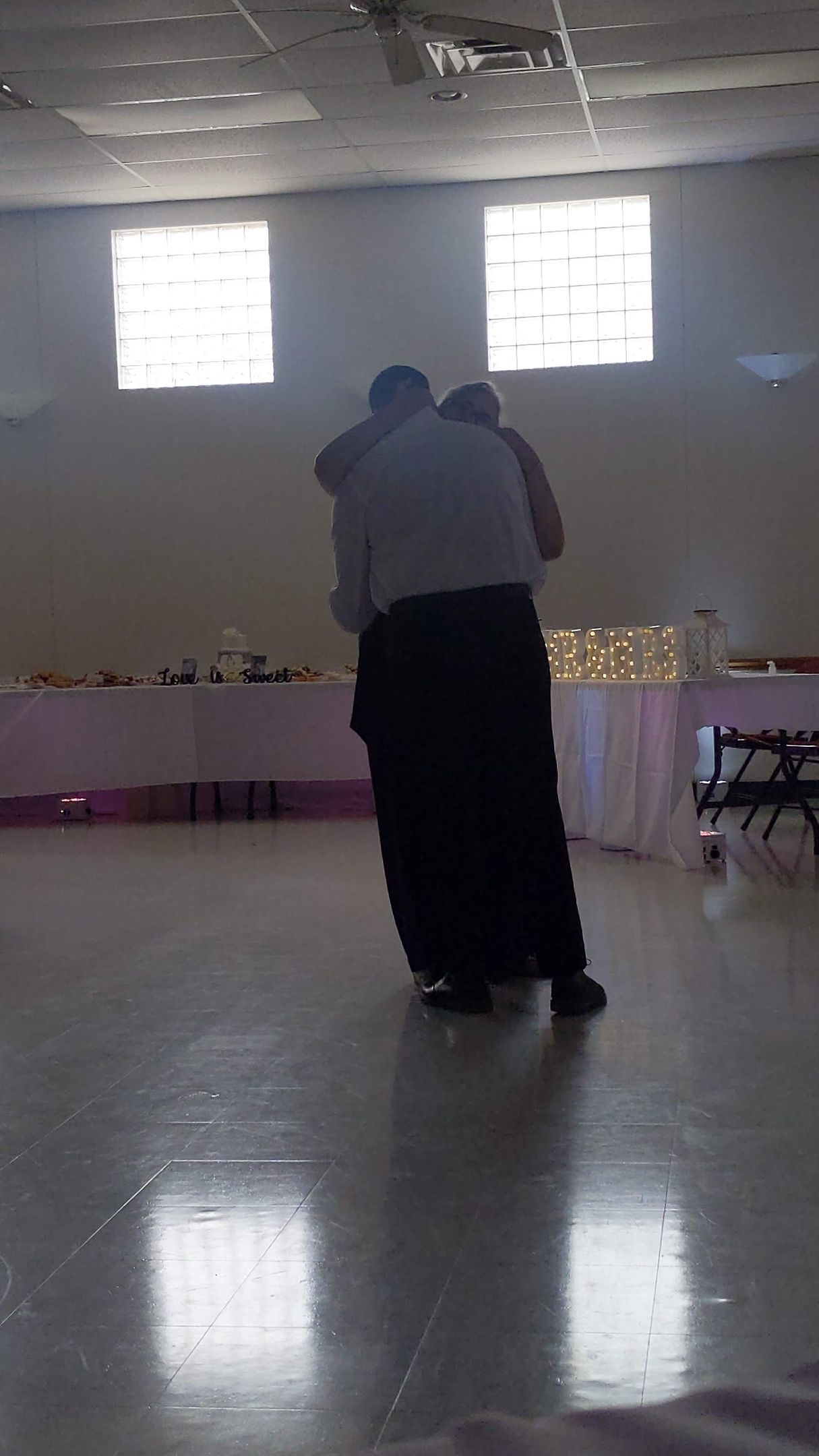 Two people embracing in a bright indoor hall with tiled floor and small windows.
