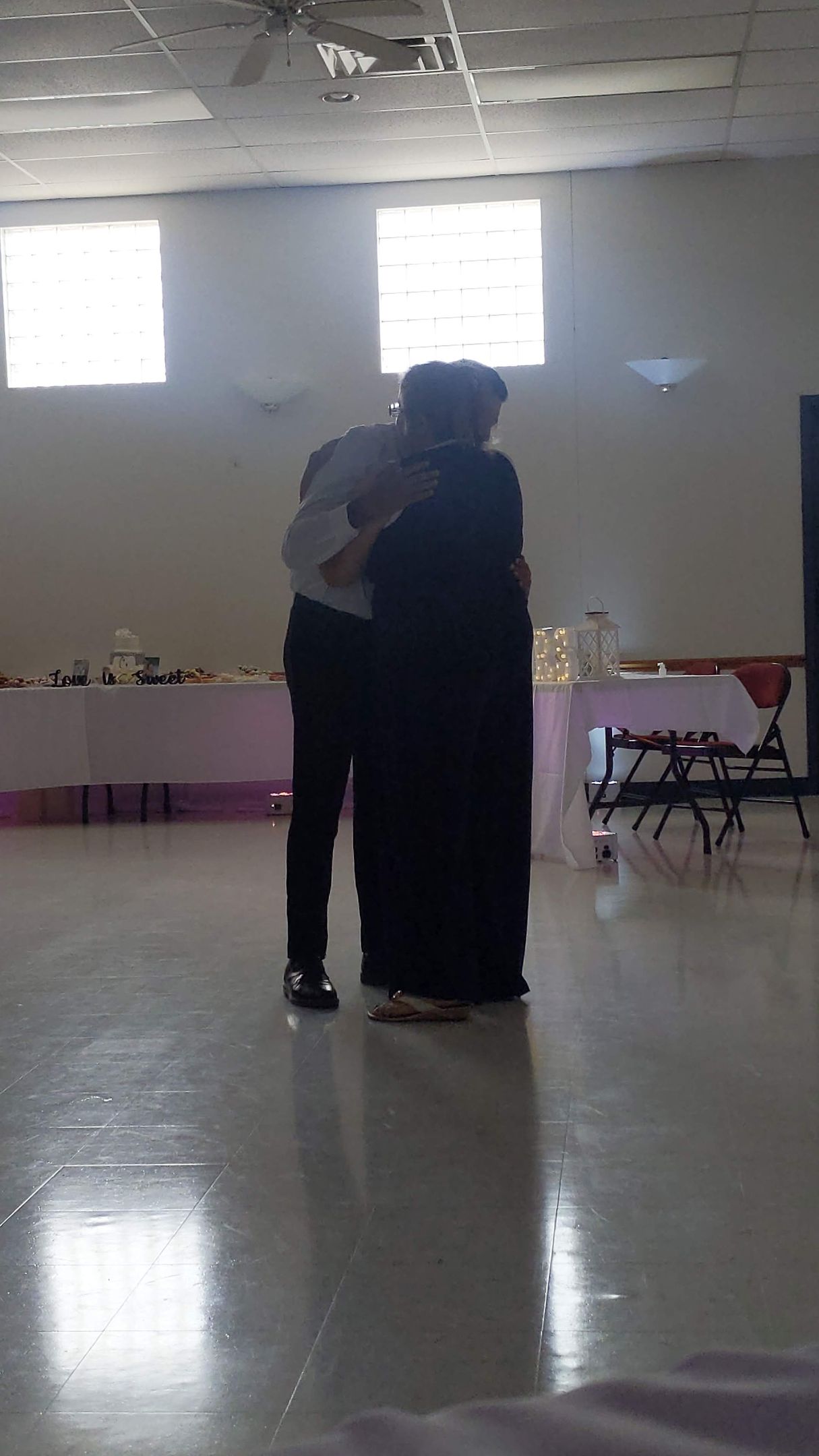 Two people embrace in a bright hall with windows and tables along the back wall.
