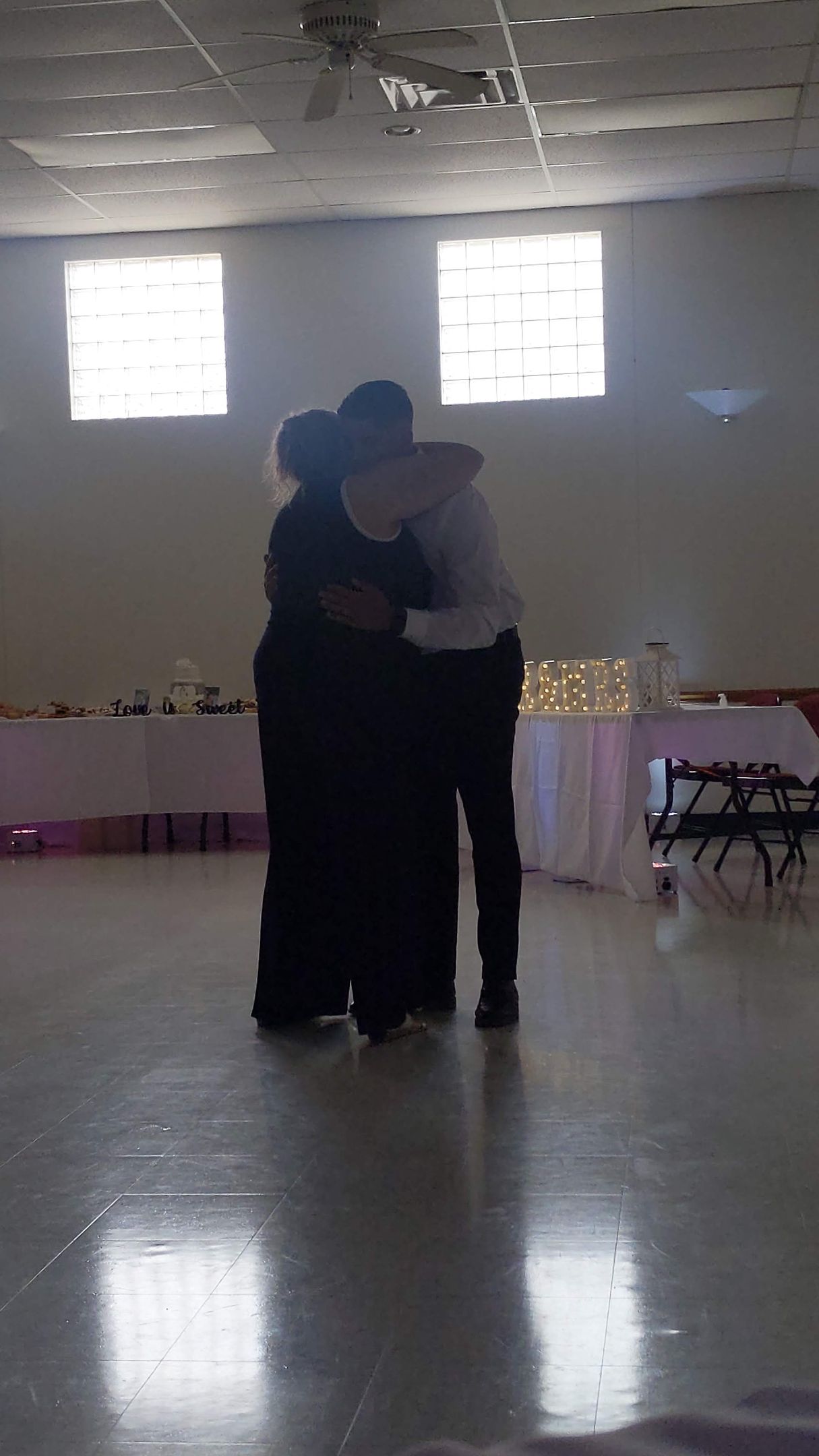 Two people embracing on a polished dance floor in a bright hall with tables along the walls.