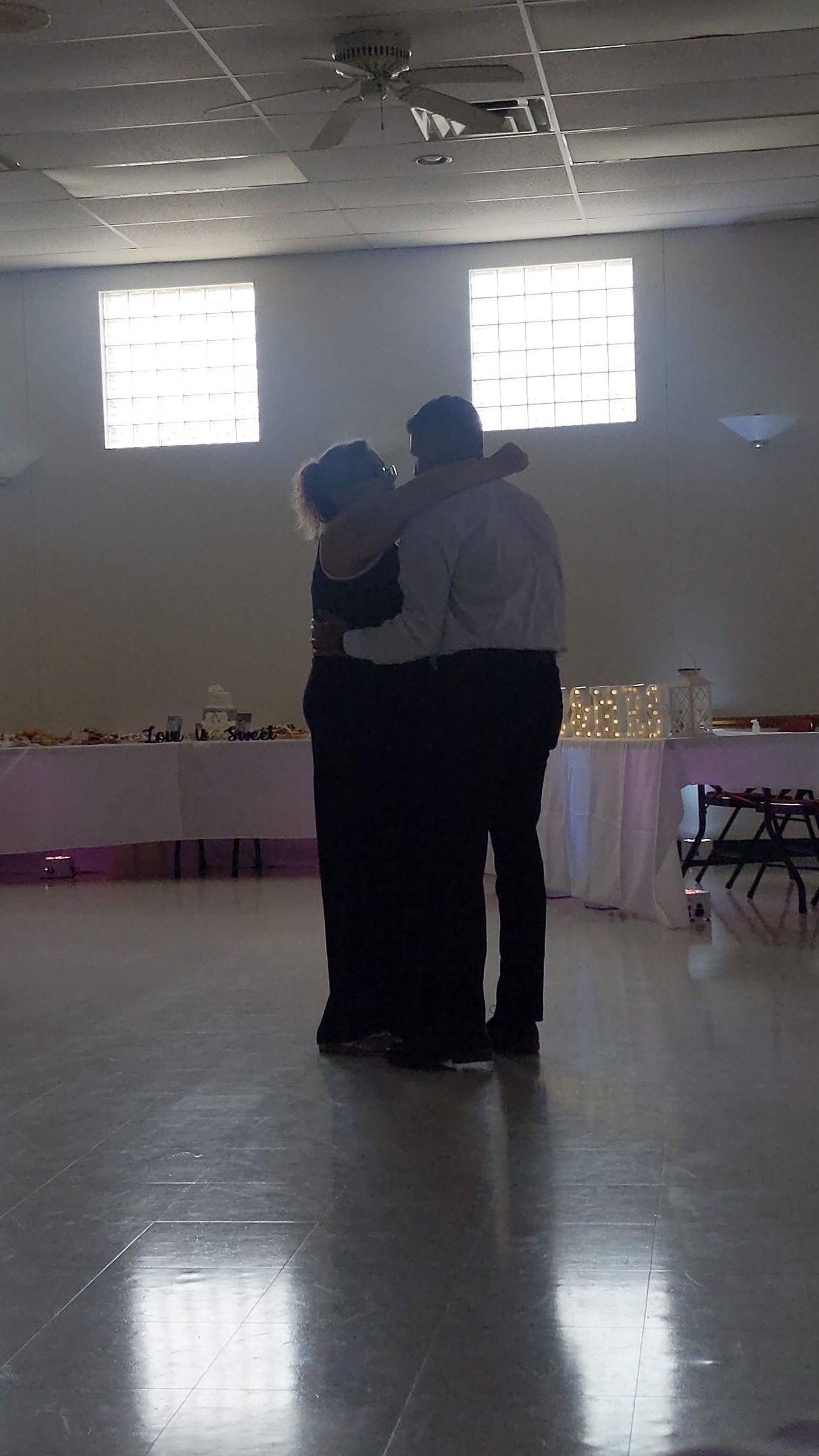 Two people embracing in a dimly lit banquet hall with tables along the back wall.