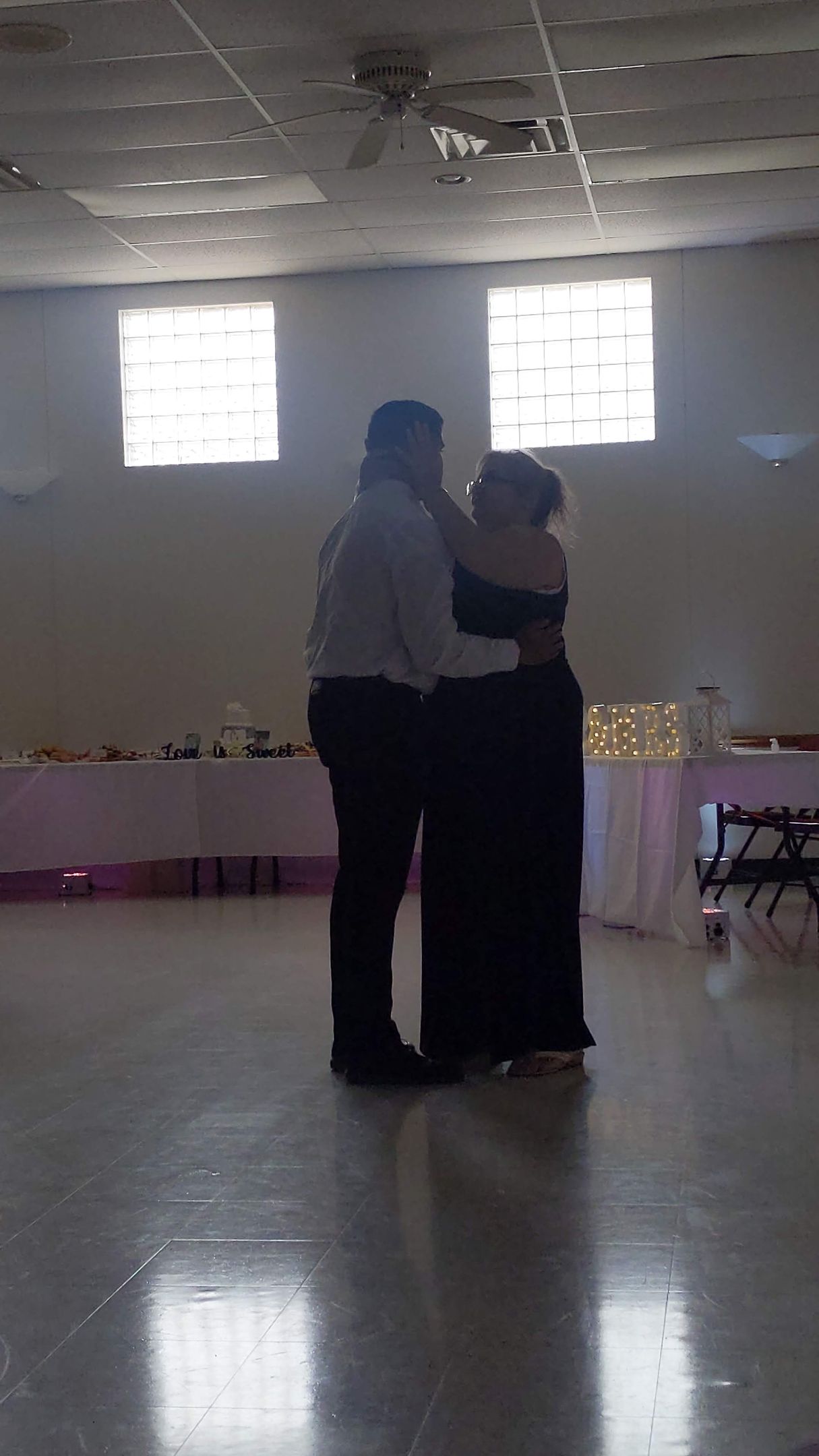 Two people dancing closely in a dimly lit hall with windows and tables along the back wall