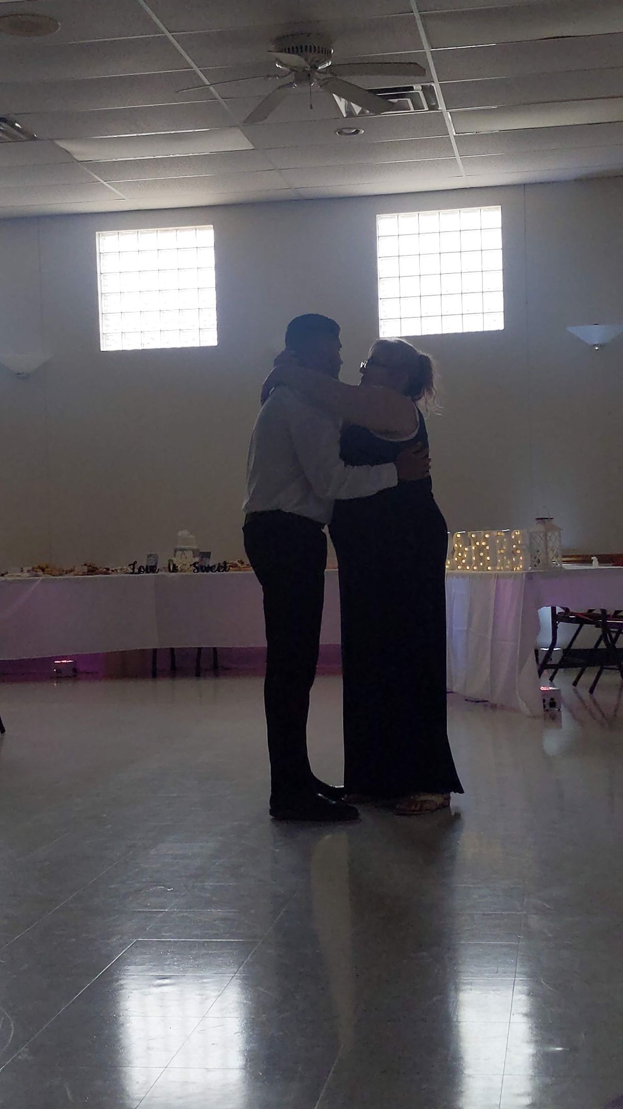 Two people dancing in a softly lit room with tables and window light in the background