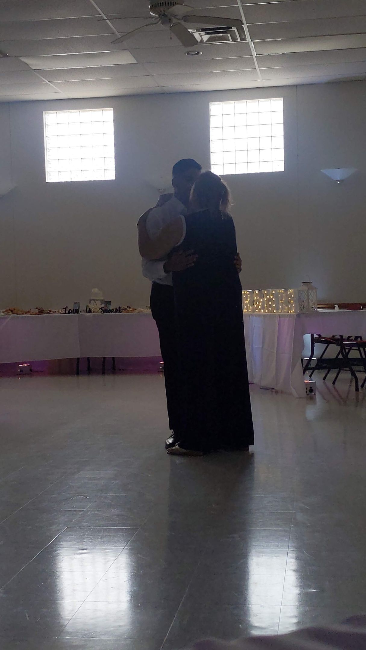 Two people embracing in a dim hall with bright windows and tables along the back wall