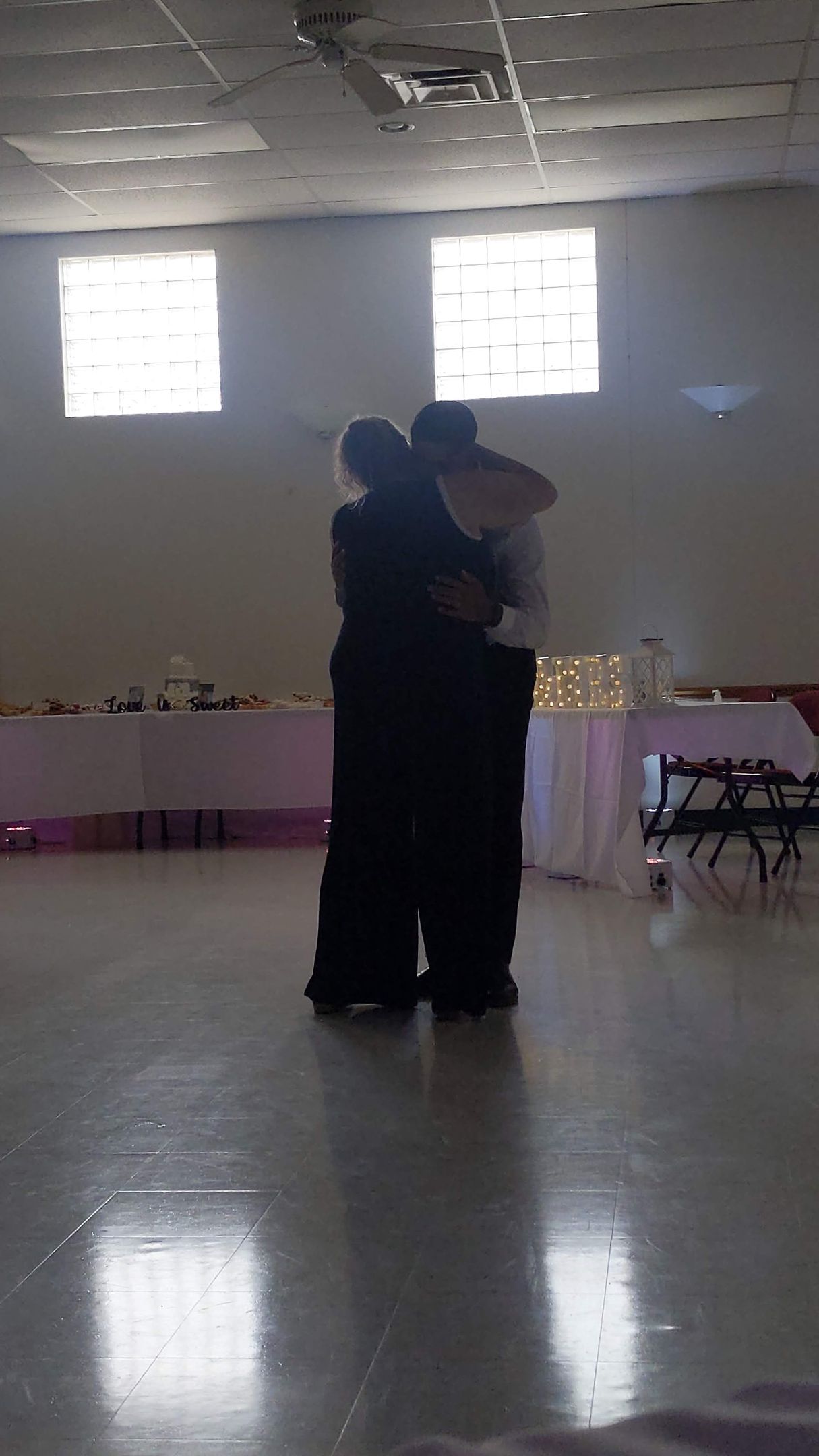 Two people embracing on a dance floor in a softly lit hall with tables and windows in the background