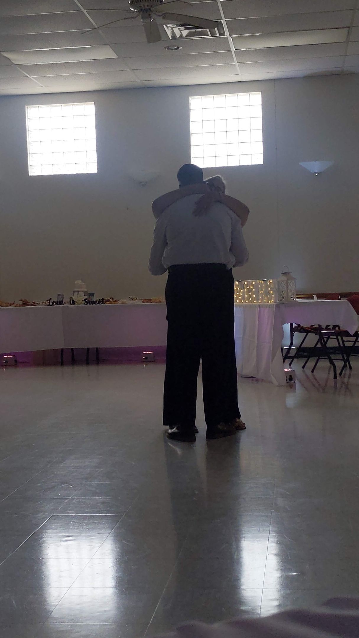 Two people embracing in a bright hall with a glossy floor and tables along the walls.