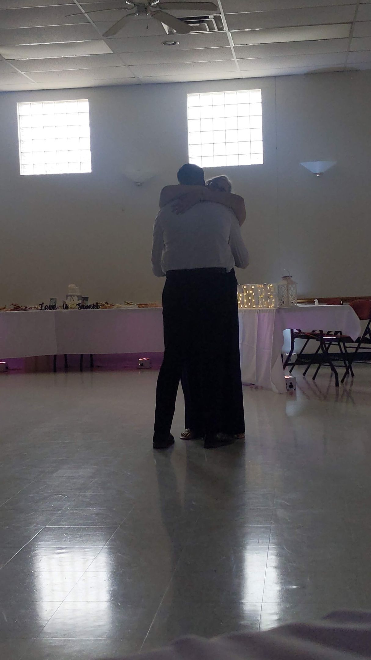 Two people embrace in a bright hall with a reflective floor and tables along the back wall.