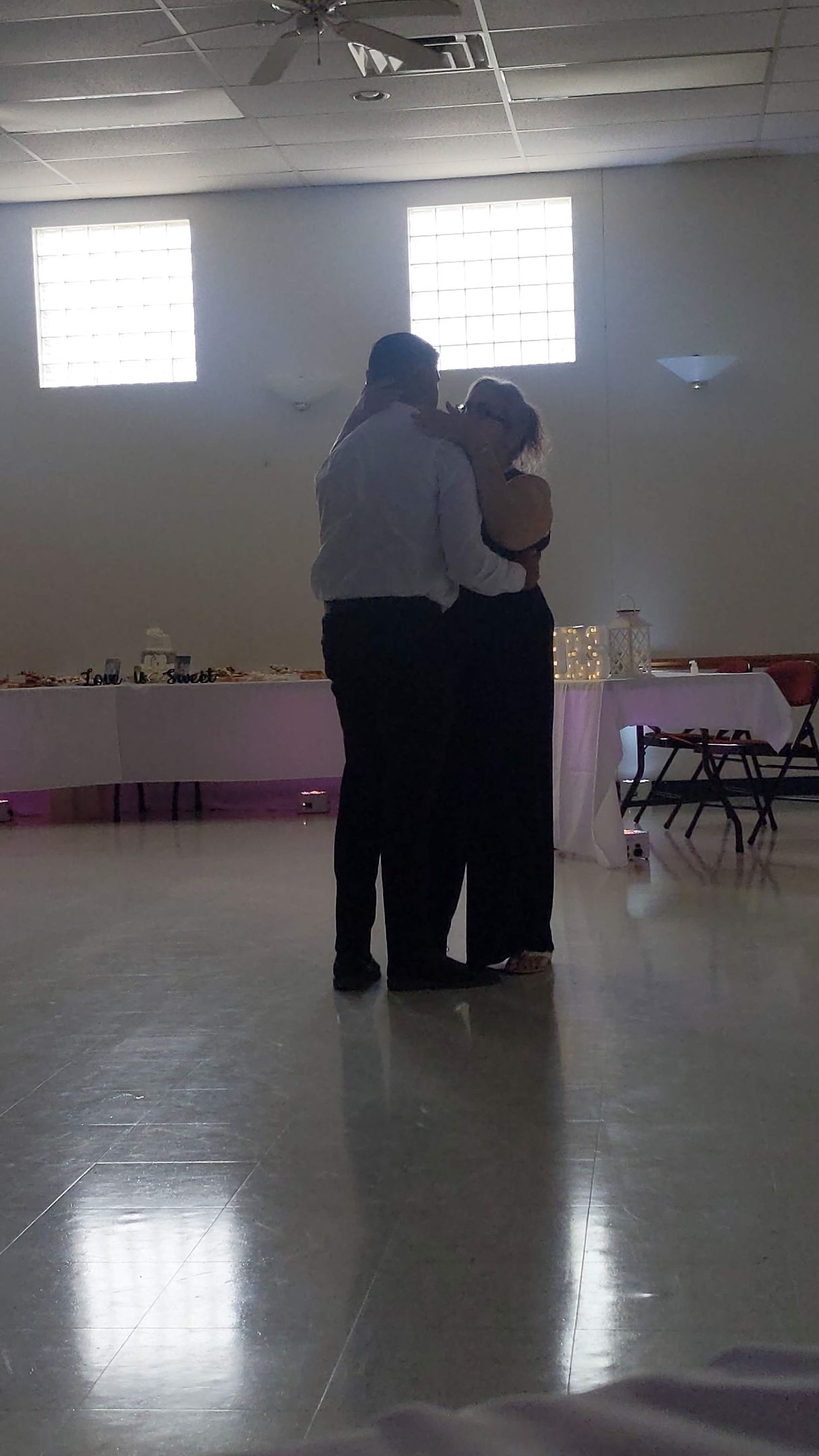 Two people dancing in a brightly lit hall with reflective floor and windows in the background