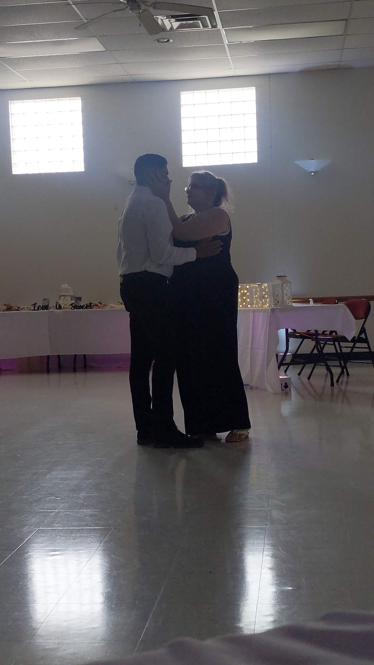 Two people dancing slowly in a bright hall with tables along the wall.