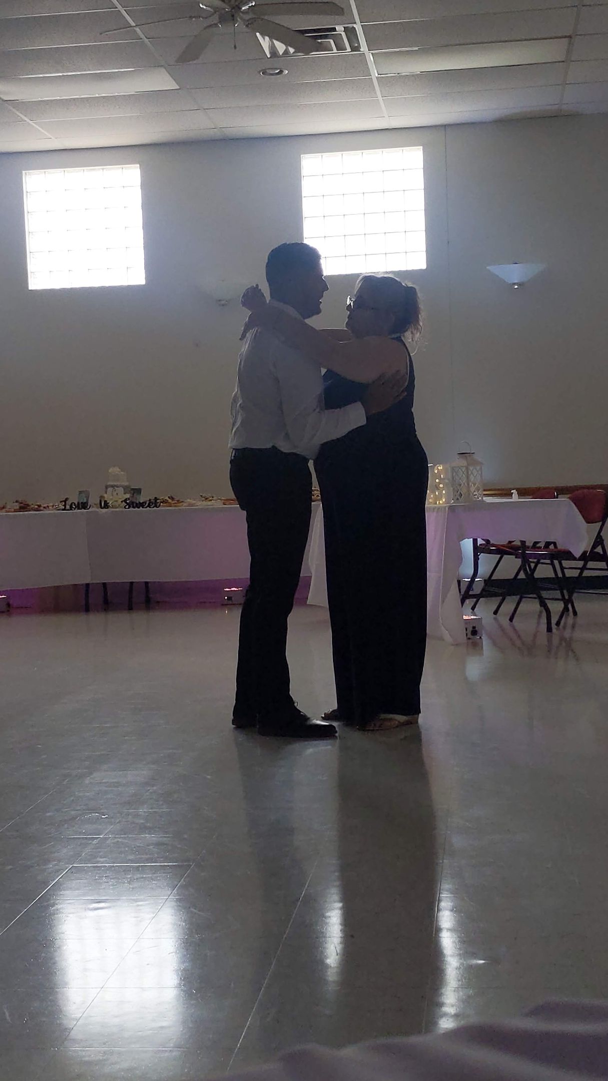 Two people dancing together in a bright room with tables and chairs along the wall.