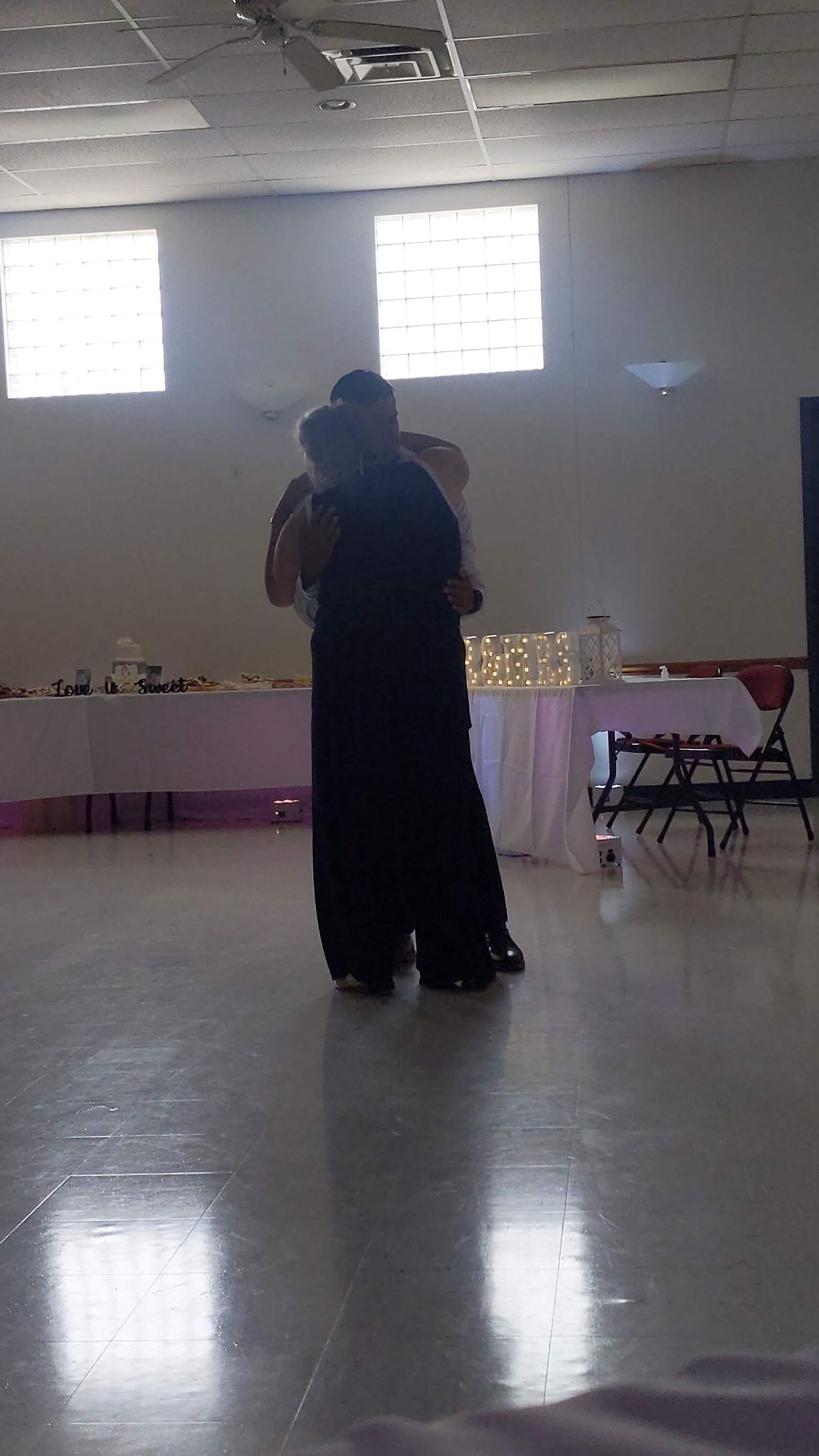 Two people embracing in a bright hall with tables along the wall