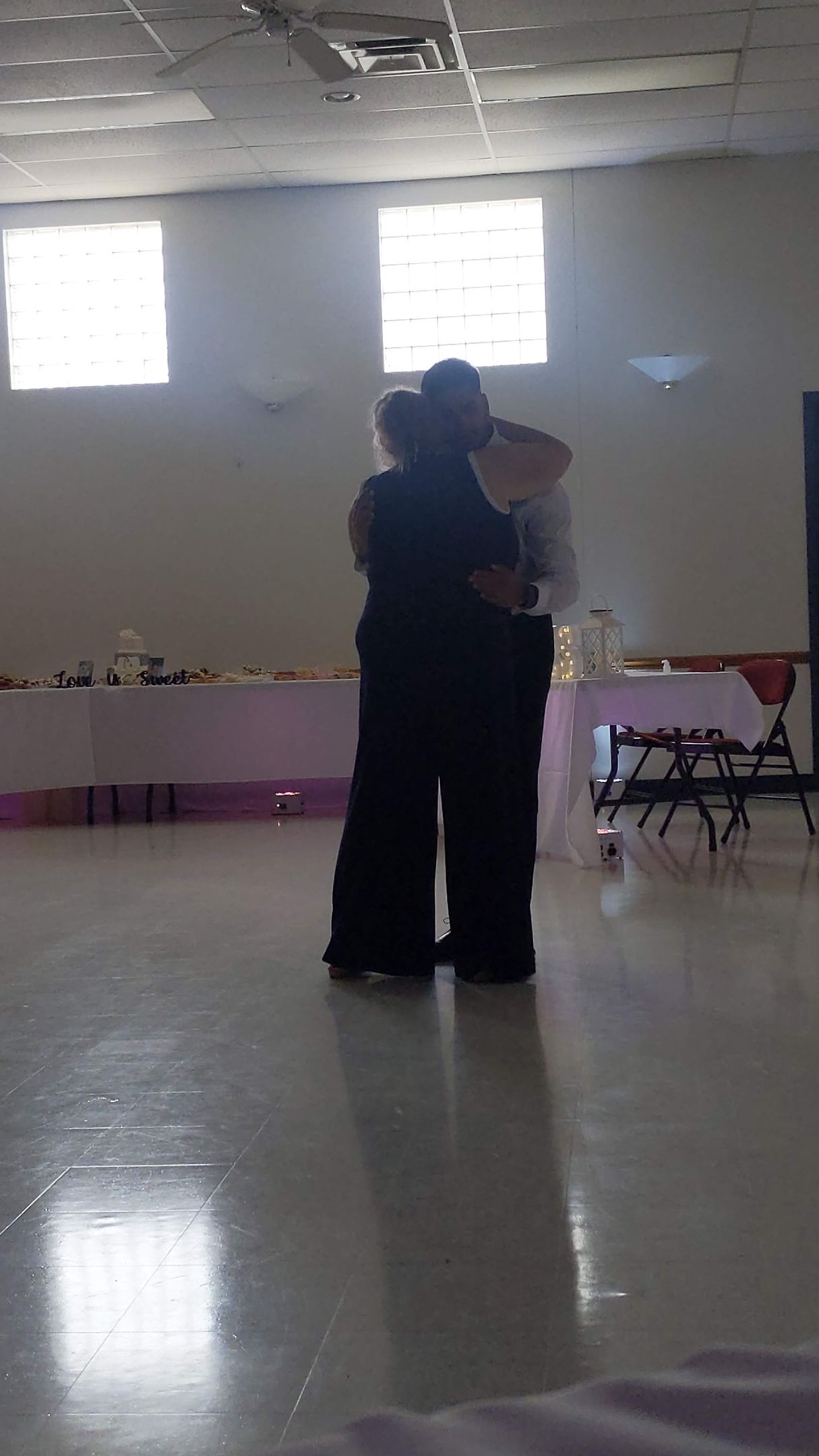 Two people dancing in a bright hall with windows and folding chairs along the wall.