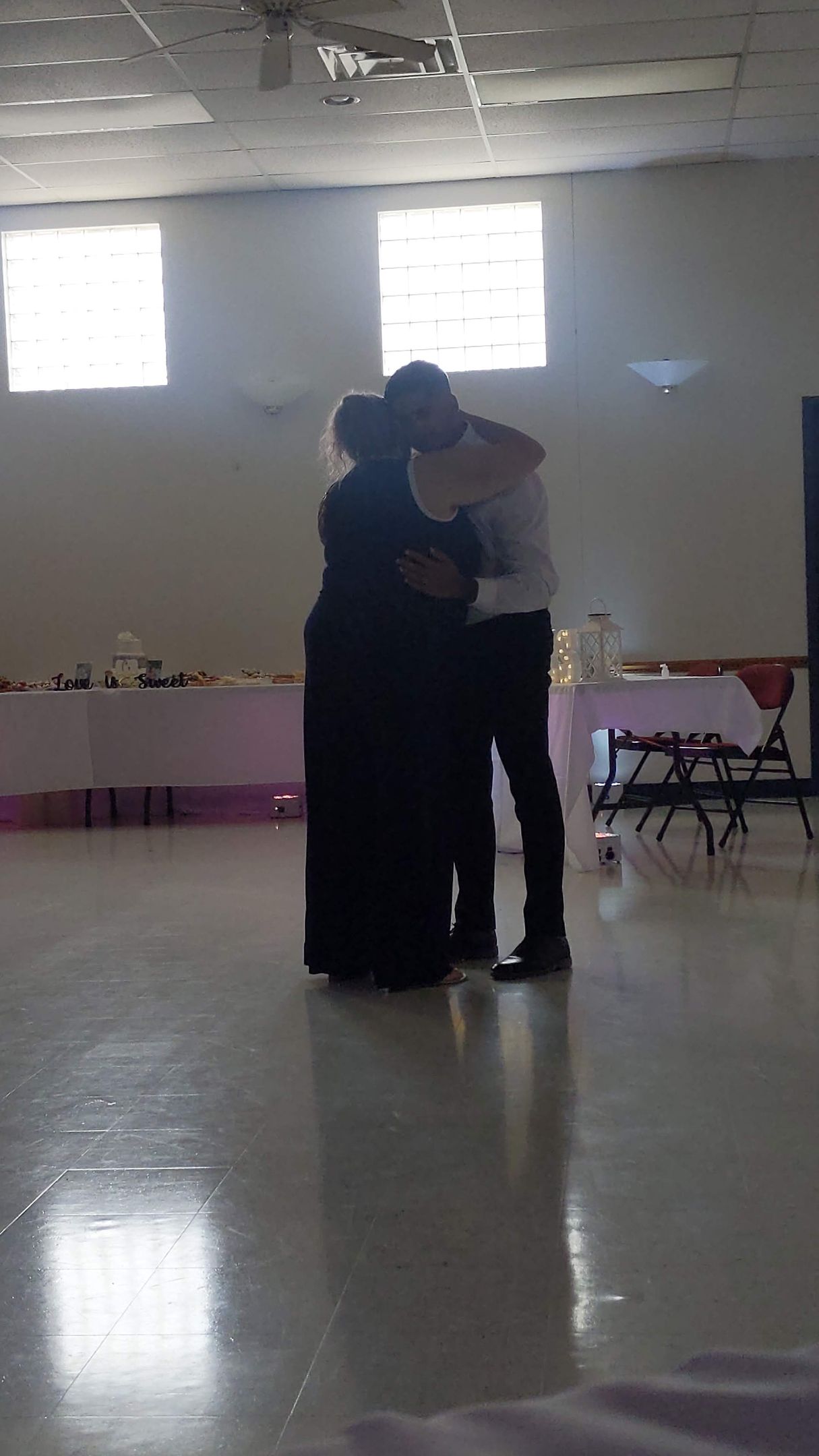 Two people embracing in a bright indoor room with windows and tables along the walls.