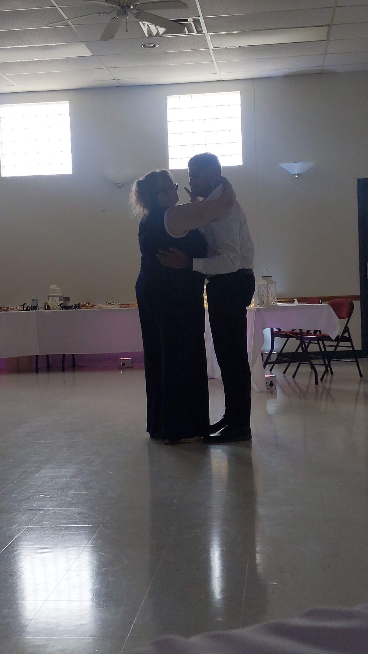 Two people embracing in a bright empty room with tiled floor and windows.