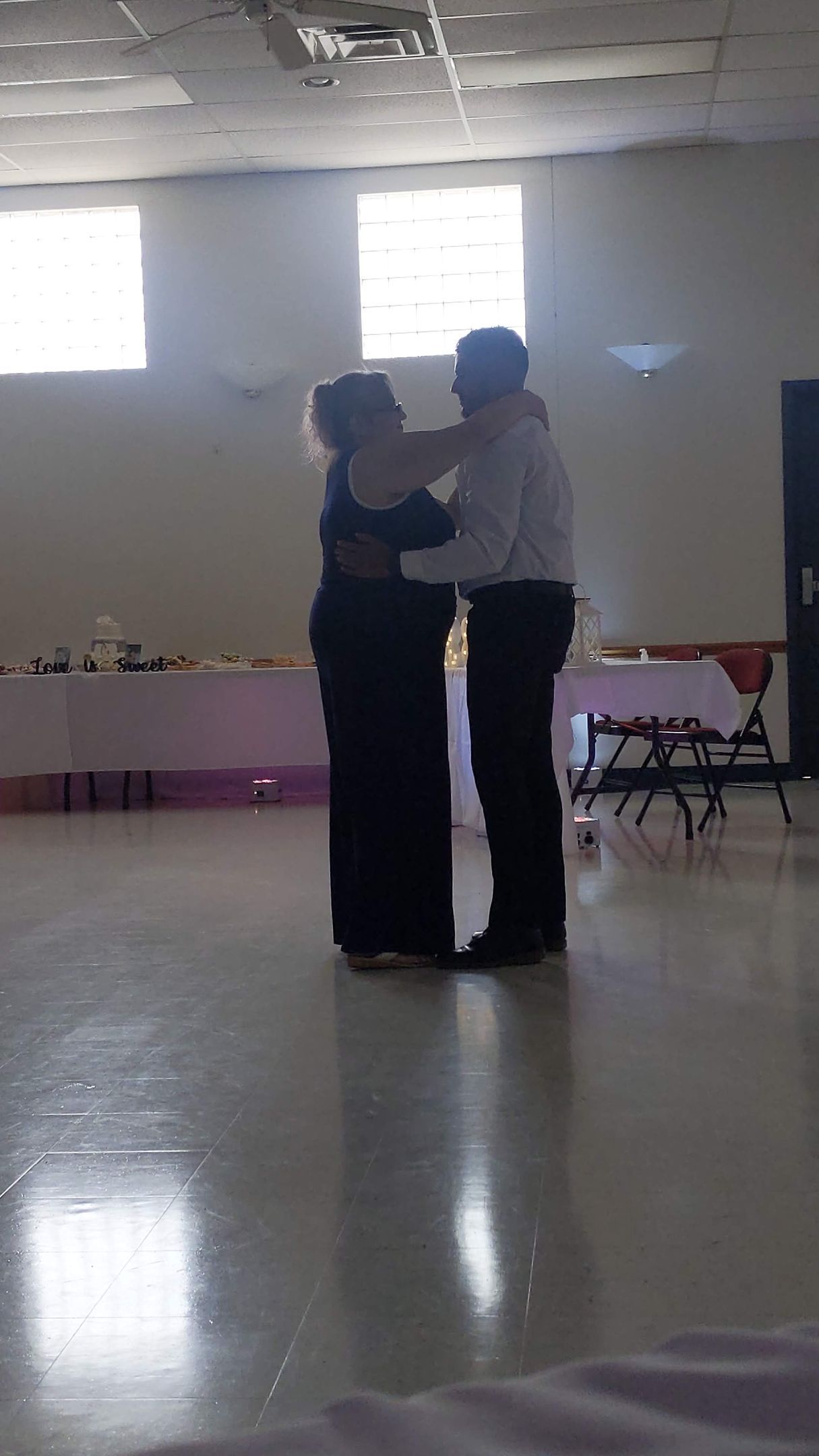 Two people dancing closely in a bright, empty room with windows and folding chairs along the wall.