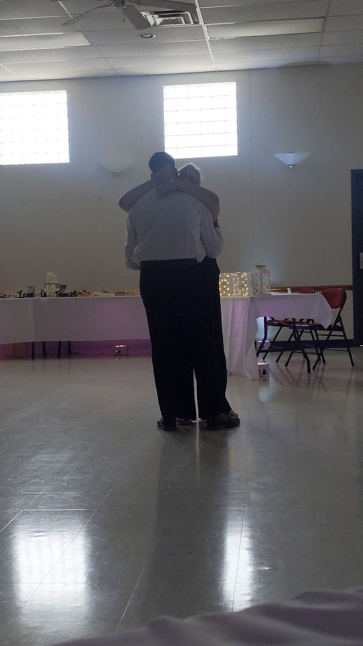 Two people embracing in a dim hall with tables and chairs along the walls