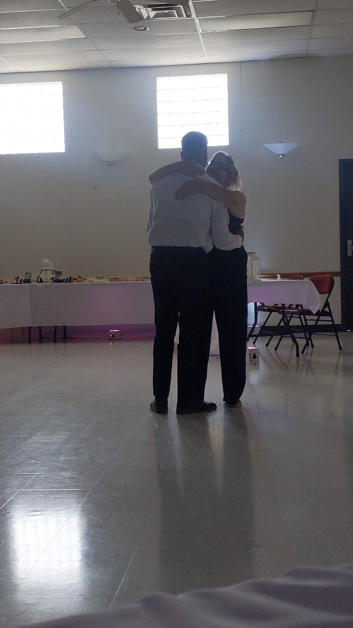 Two people embracing in a bright, empty hall with chairs along the wall.