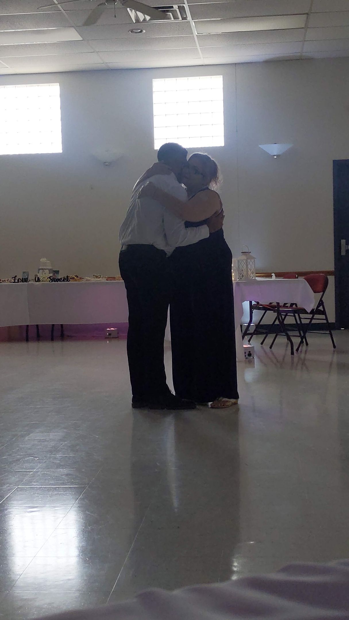 Two people embracing in a bright, empty hall with polished floor and windows.