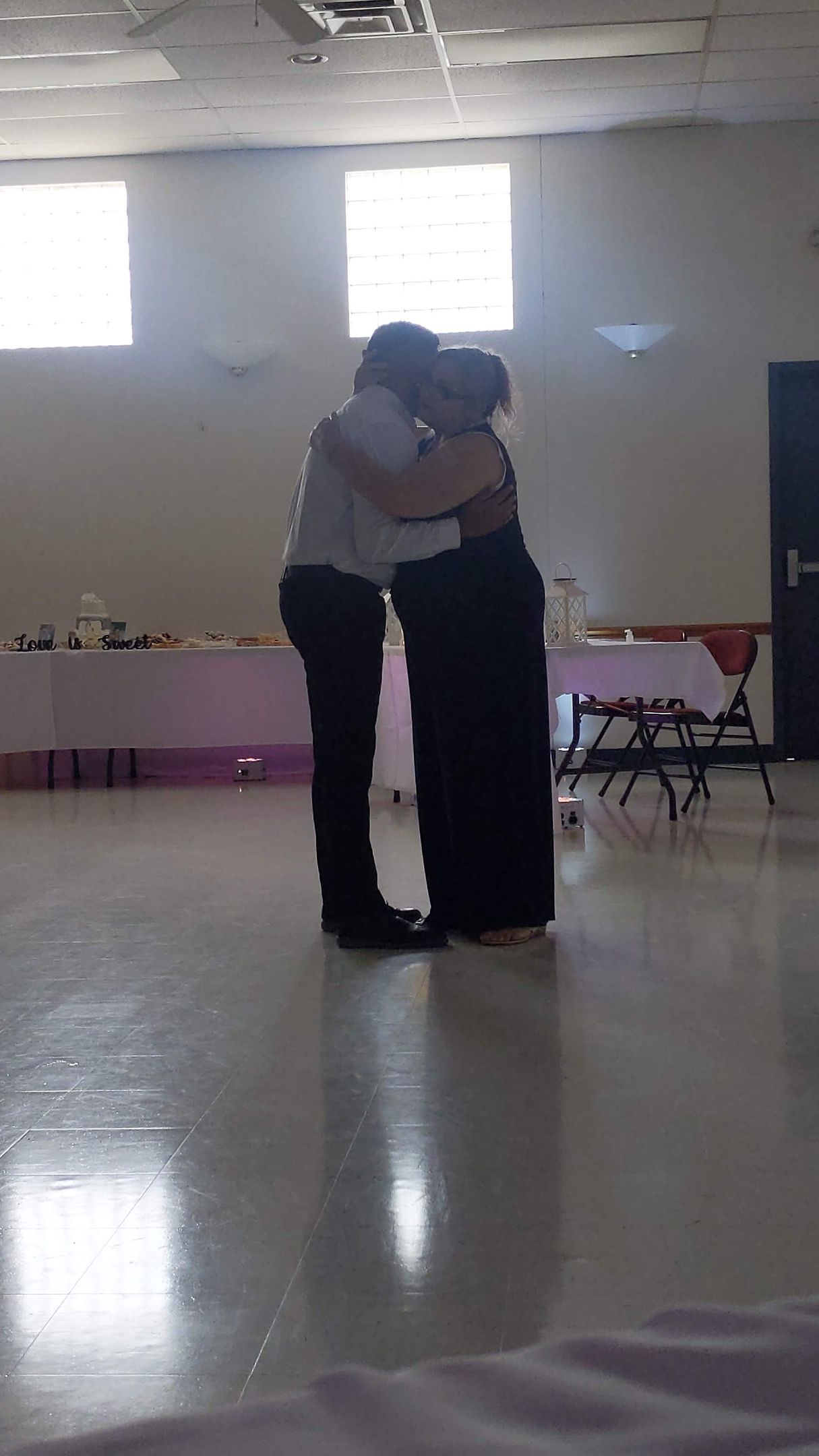 Two people embracing in a bright hall with tables along the back wall.