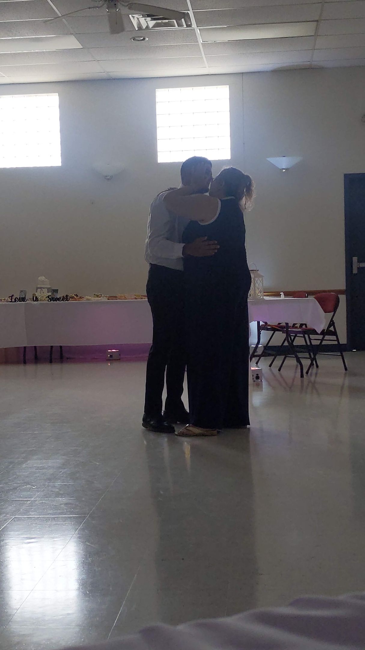 Two people embrace in a bright gymnasium with polished floors and tables along the wall.