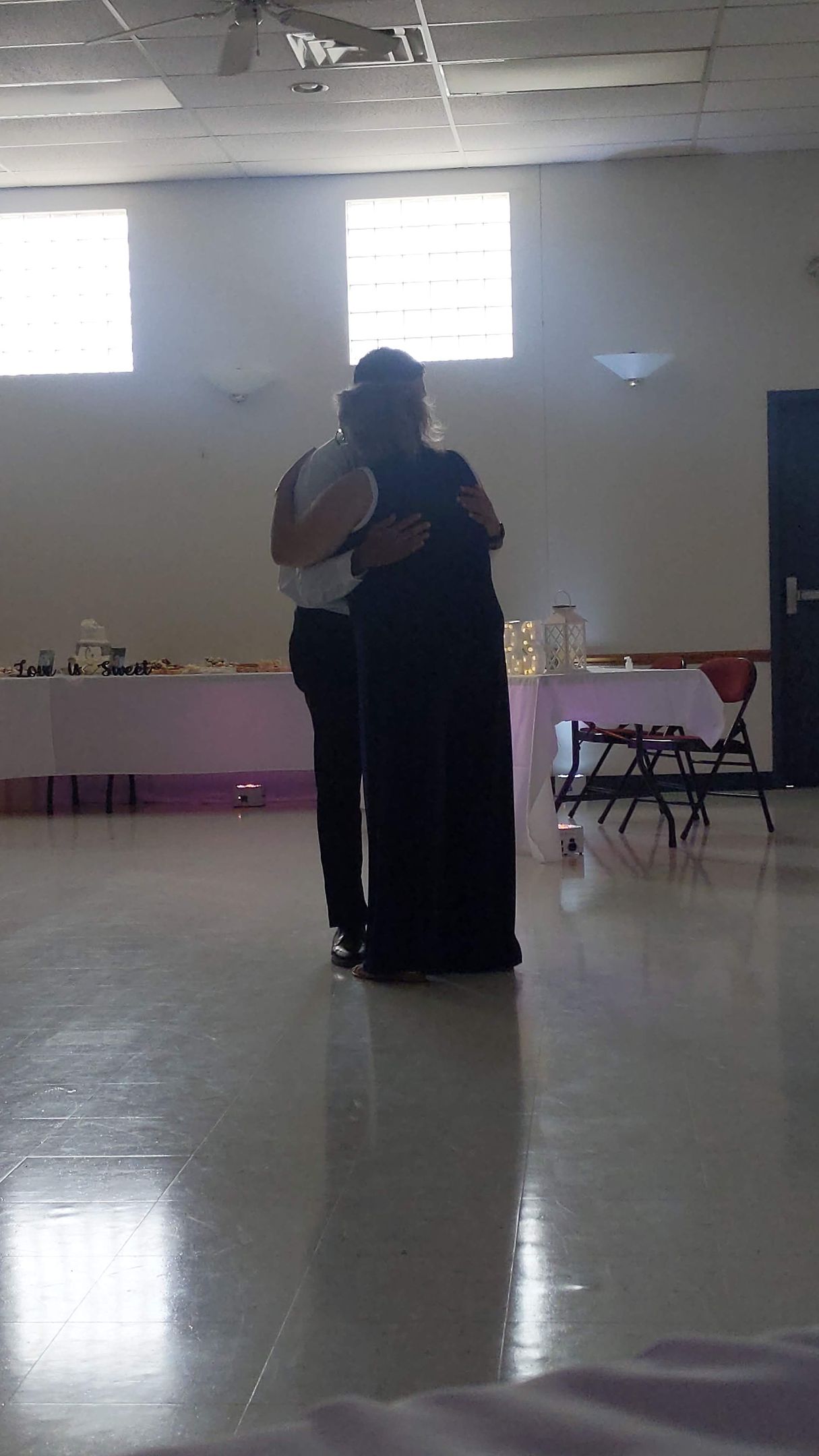 Two people embracing in a bright hall with tables along the back wall.