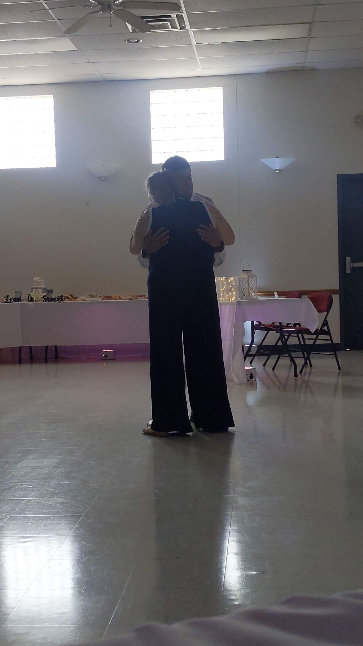 Person standing in a bright hall, centered beneath two windows, holding a phone or camera.