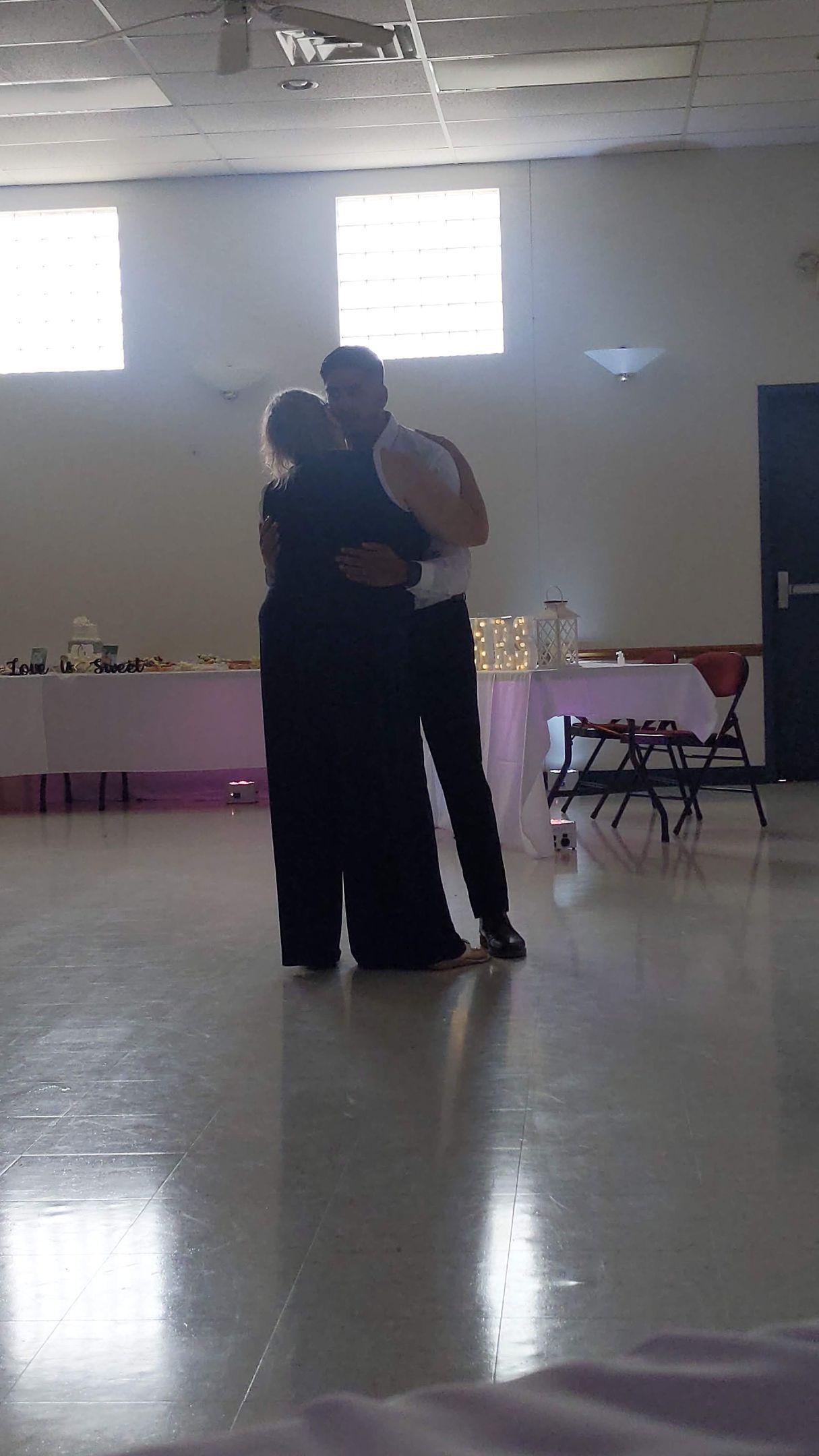 Two people embracing on a dance floor in a bright hall with tables along the wall.