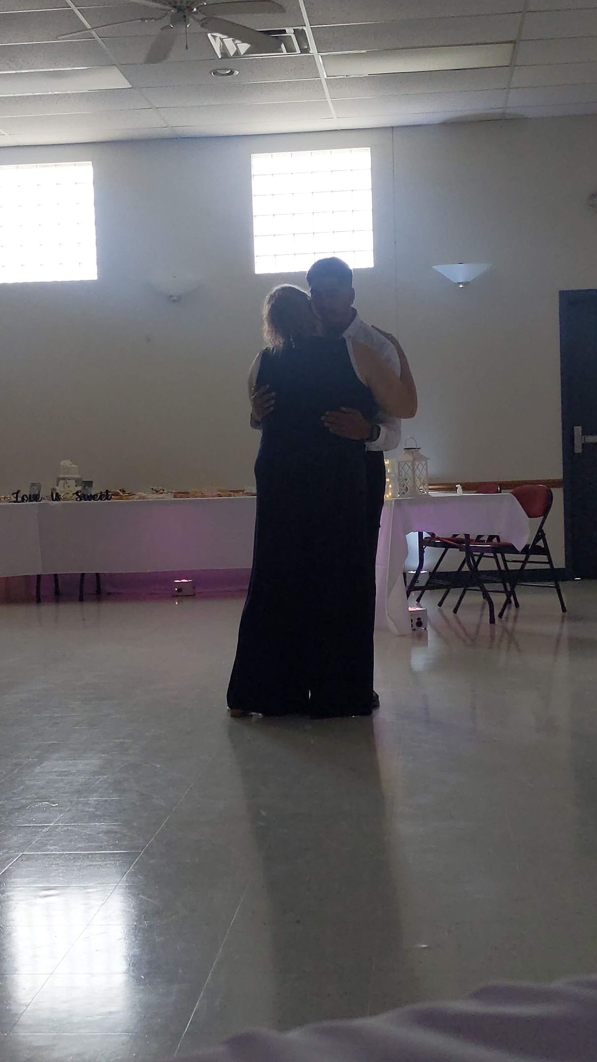 Two people embracing in a bright indoor hall with tiled floor and high windows.