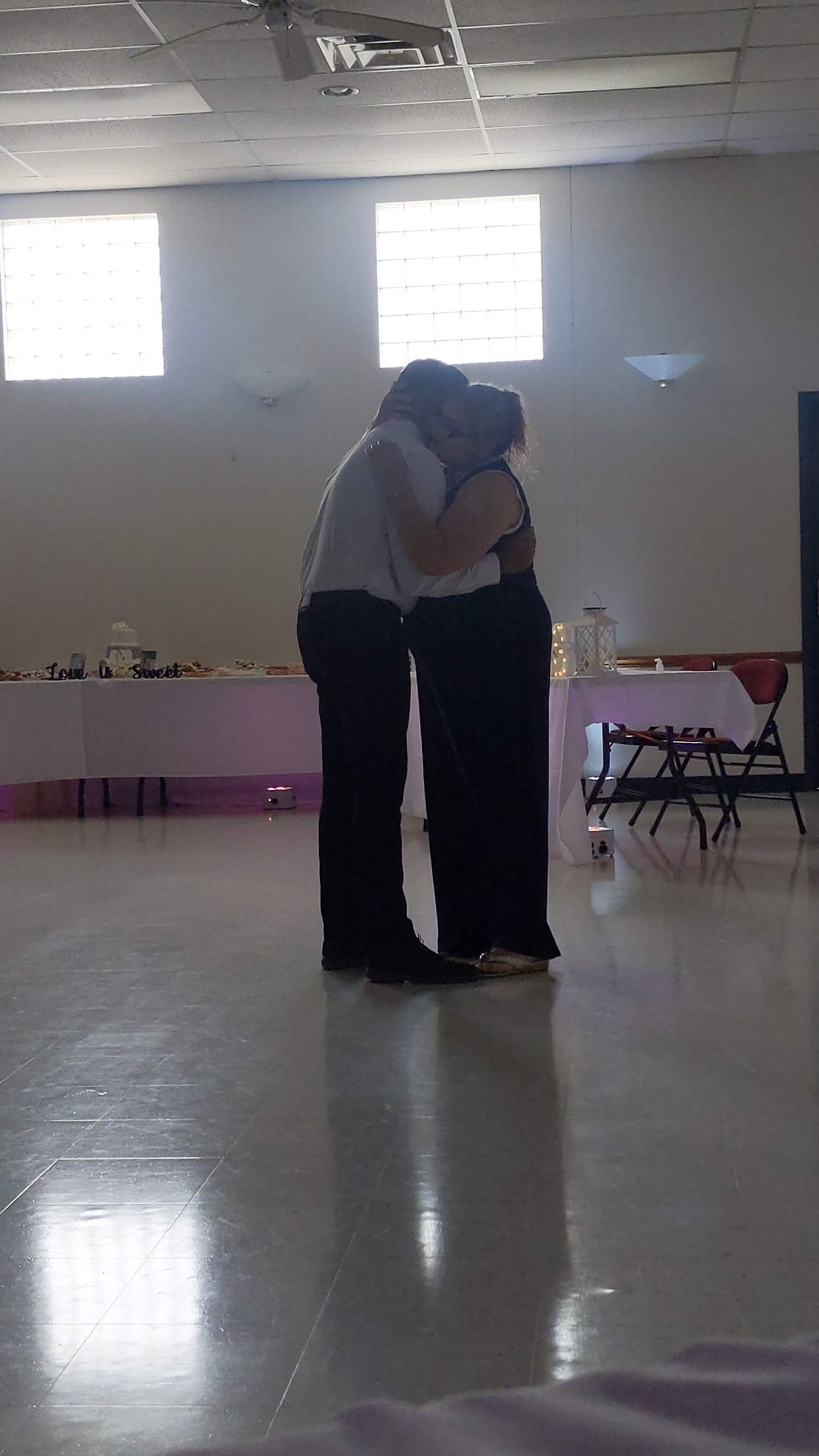 Two people embracing in a bright hall with a reflective floor and tables along the wall