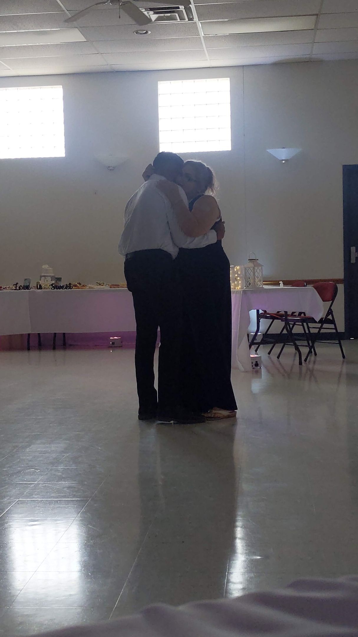 Two people embracing in a bright room with windows and a reflective floor.