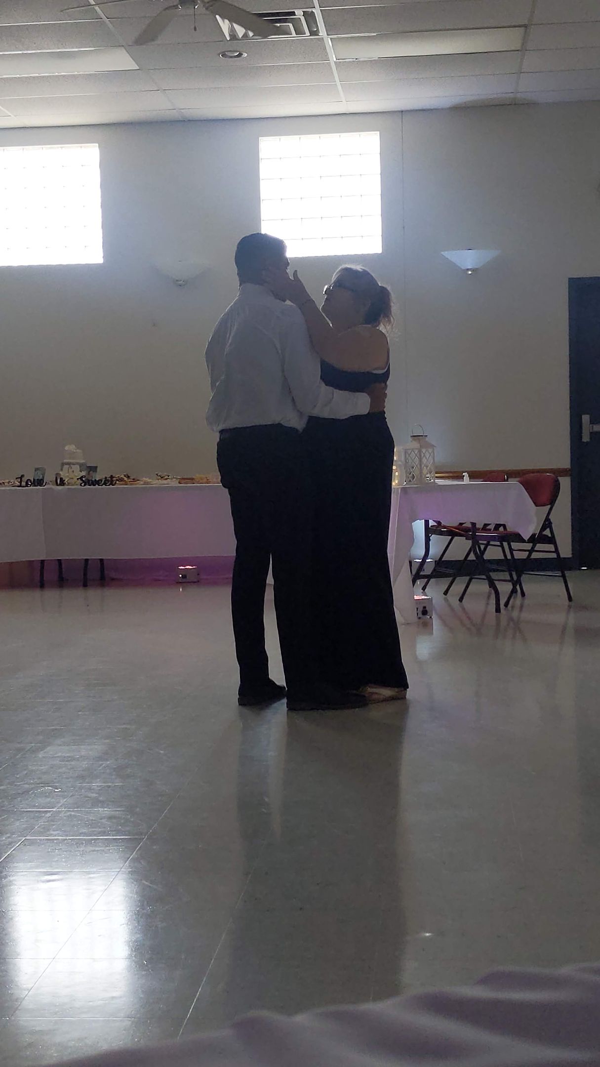 Two people dancing in a bright hall with tables along the wall.