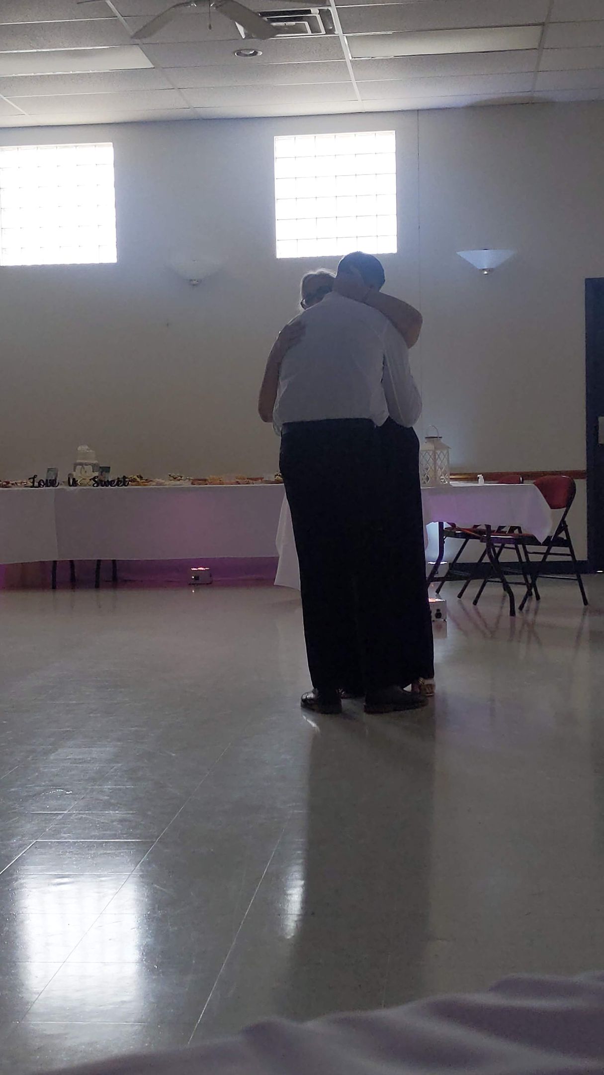 Two people embracing in a bright hall with tables along the back wall.