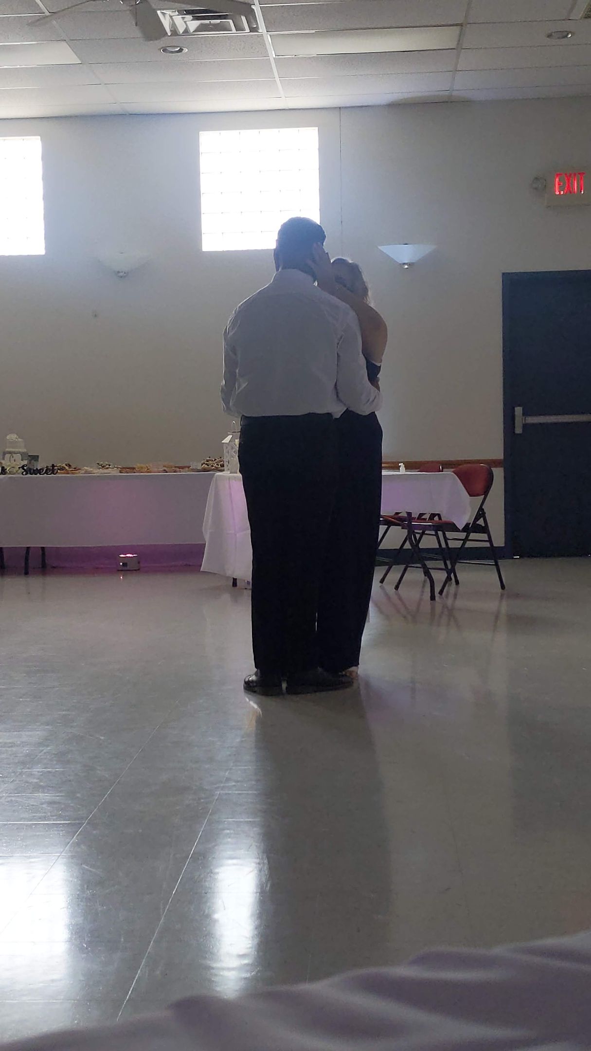 Two people embracing in a bright hall with a polished floor and tables along the wall.