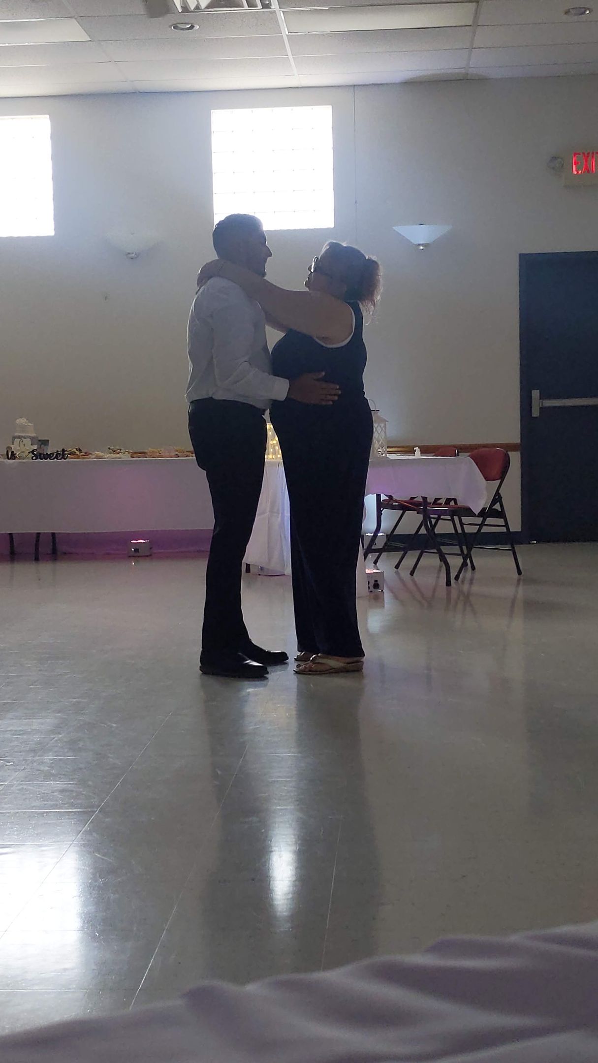 Two people dancing in a bright community hall with tables and chairs in the background