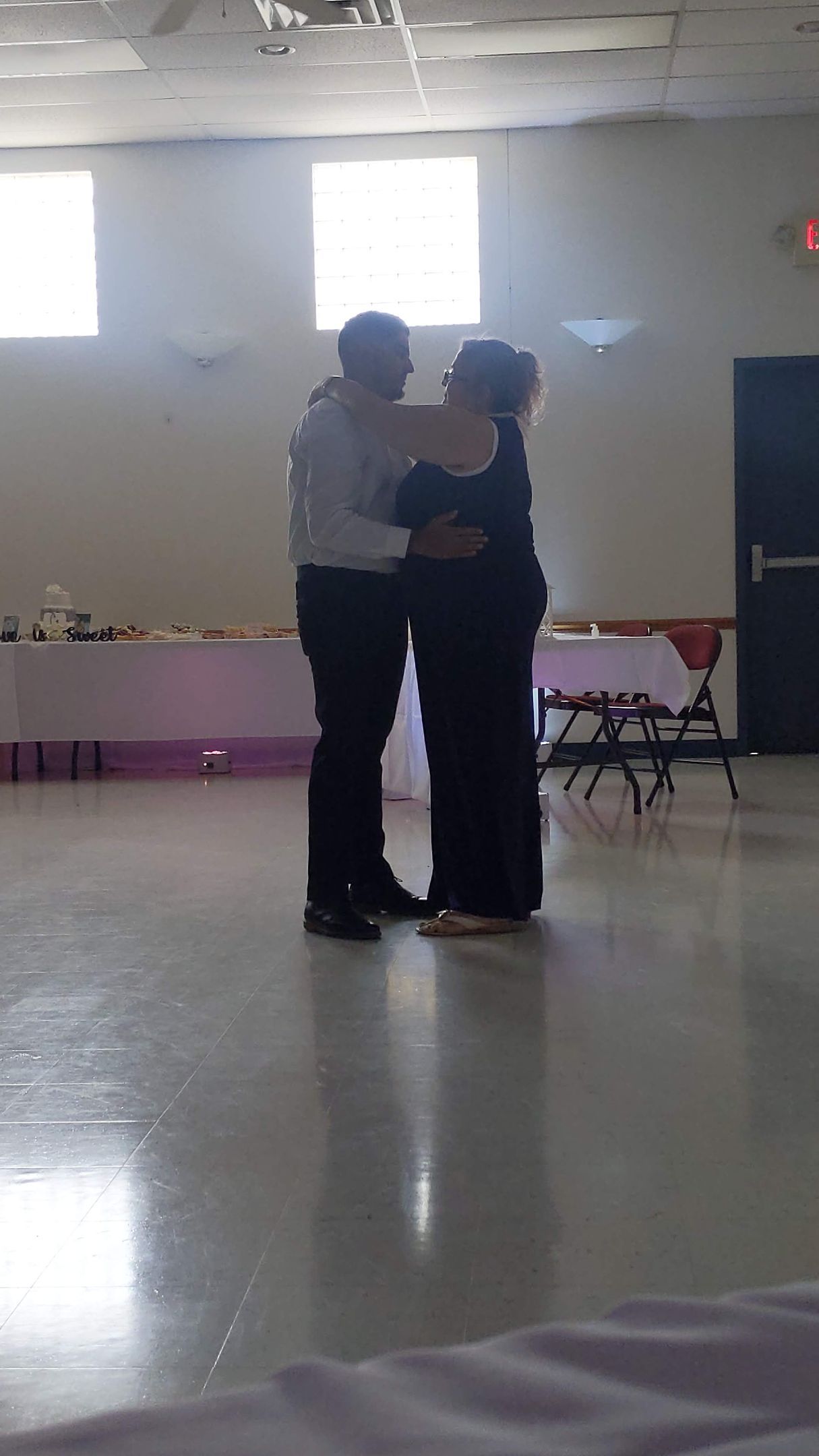 Two people slow dancing in a bright community hall with polished floor and tables along the wall