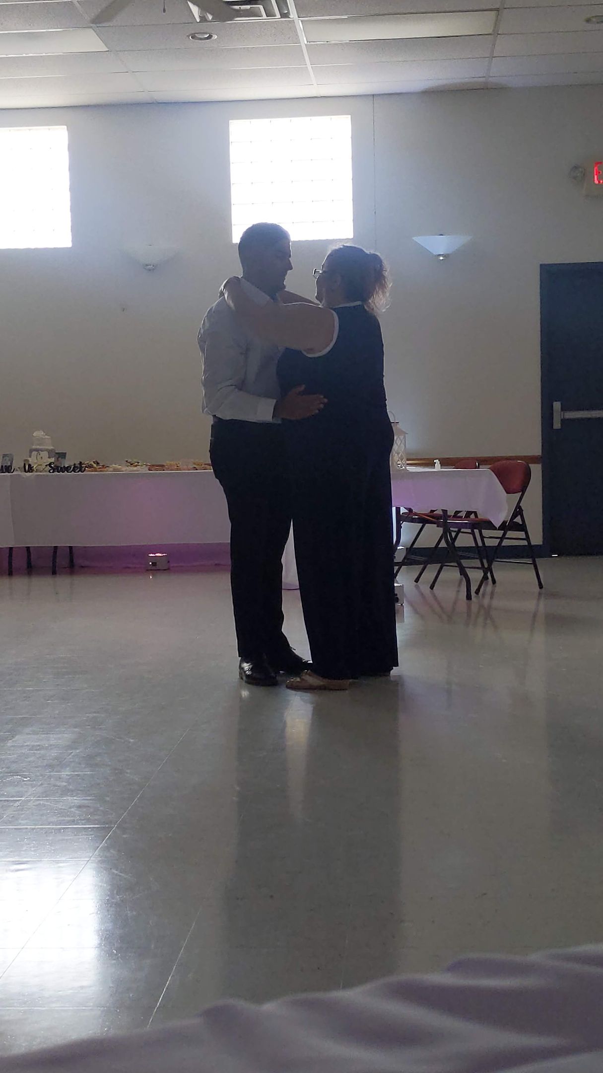 Two people embrace in a bright indoor room with tables and chairs along the wall.