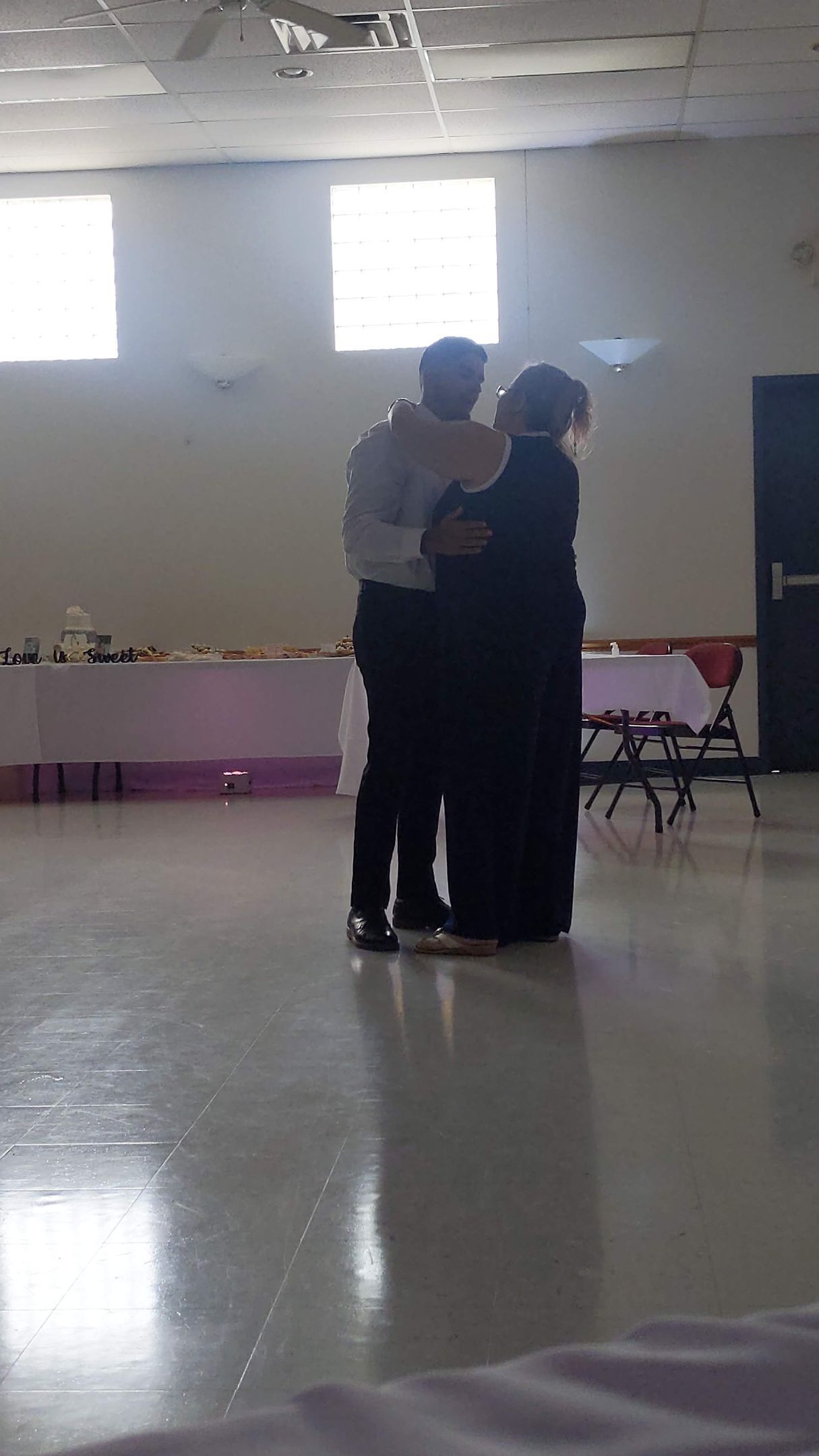 Two people slow dancing in a hall with tables and bright windows in the background