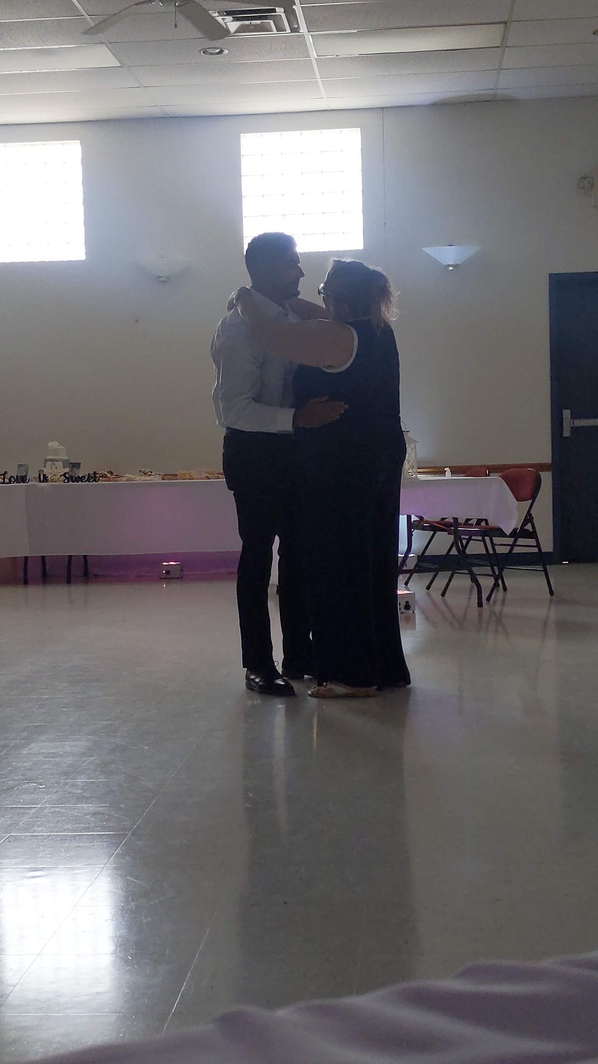 Two people embracing in a bright room with folded chairs and tables along the walls.