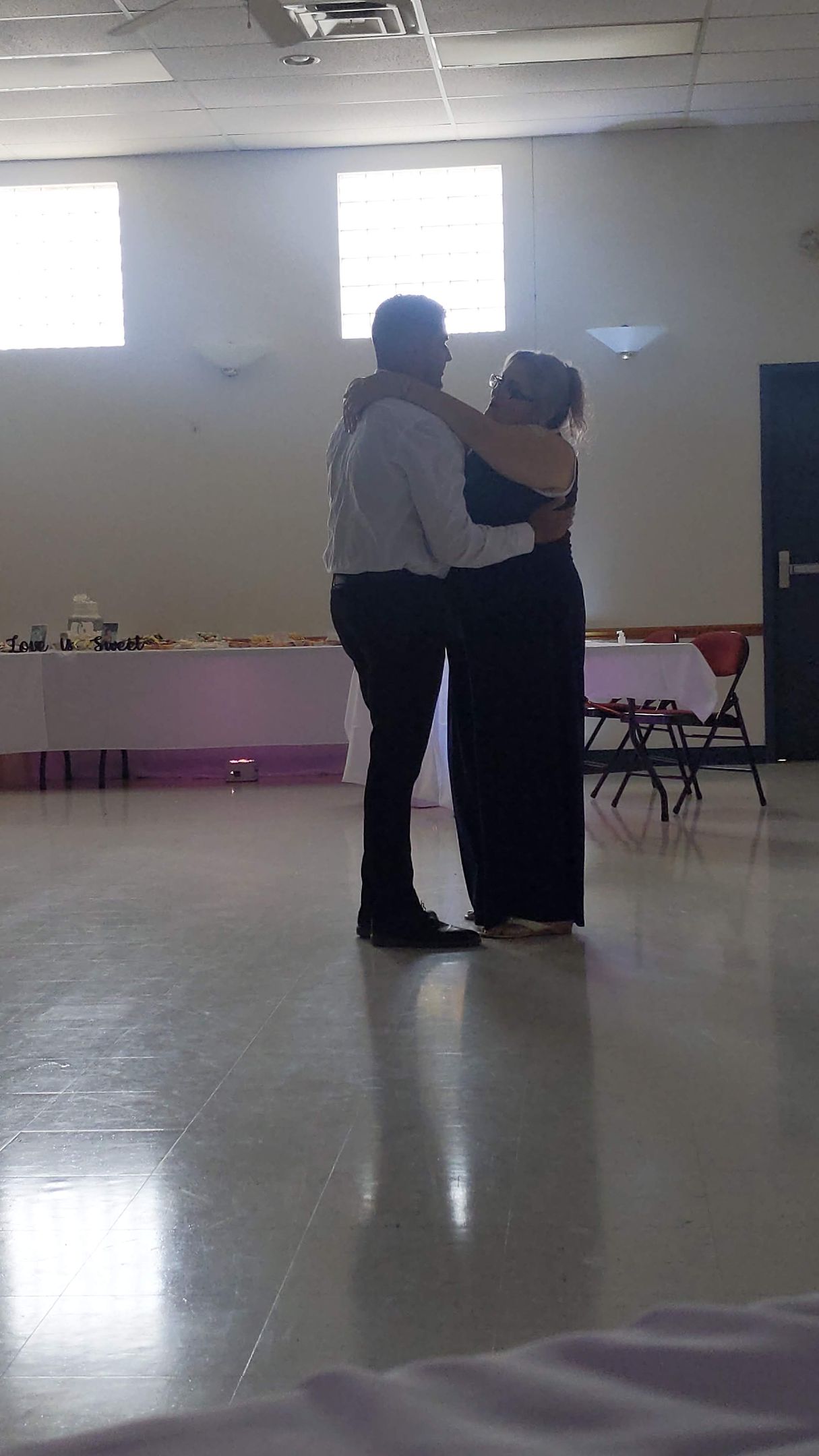 Two people embracing on a dance floor in a bright hall with tables along the wall