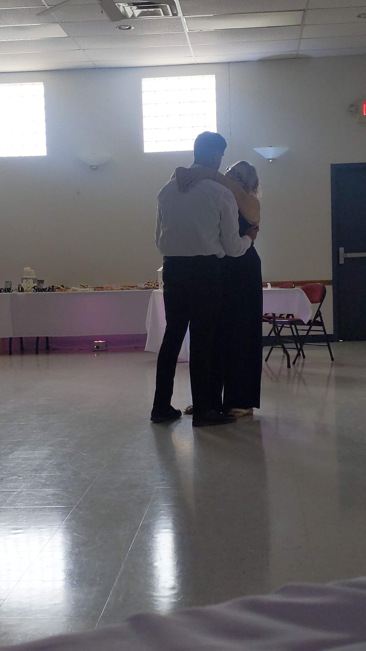 Two people embracing in a bright hall with tables and windows in the background
