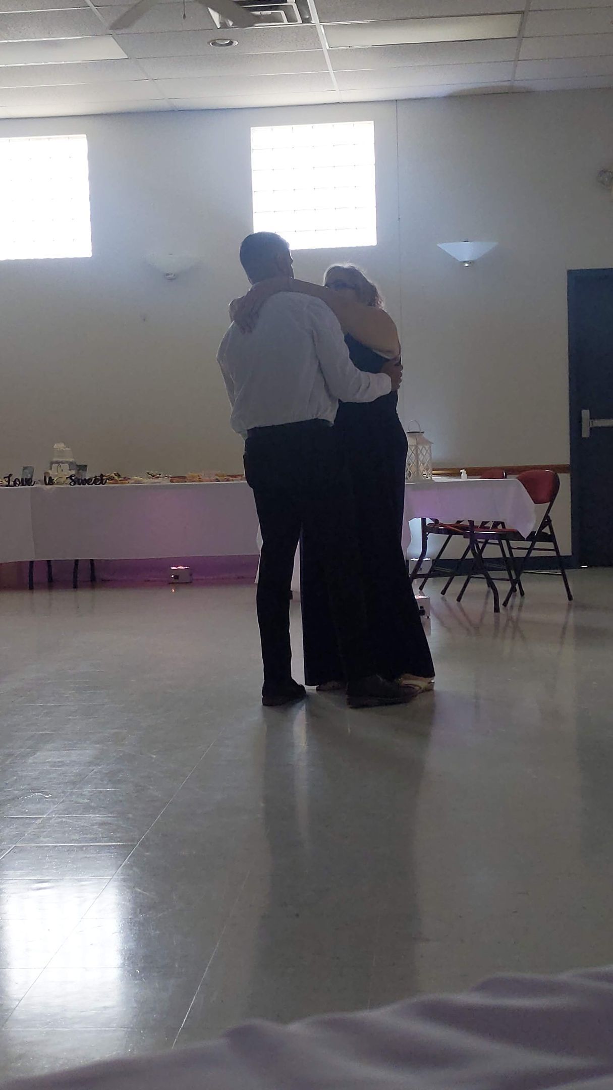 Two people dancing in a bright hall with tables along the walls.