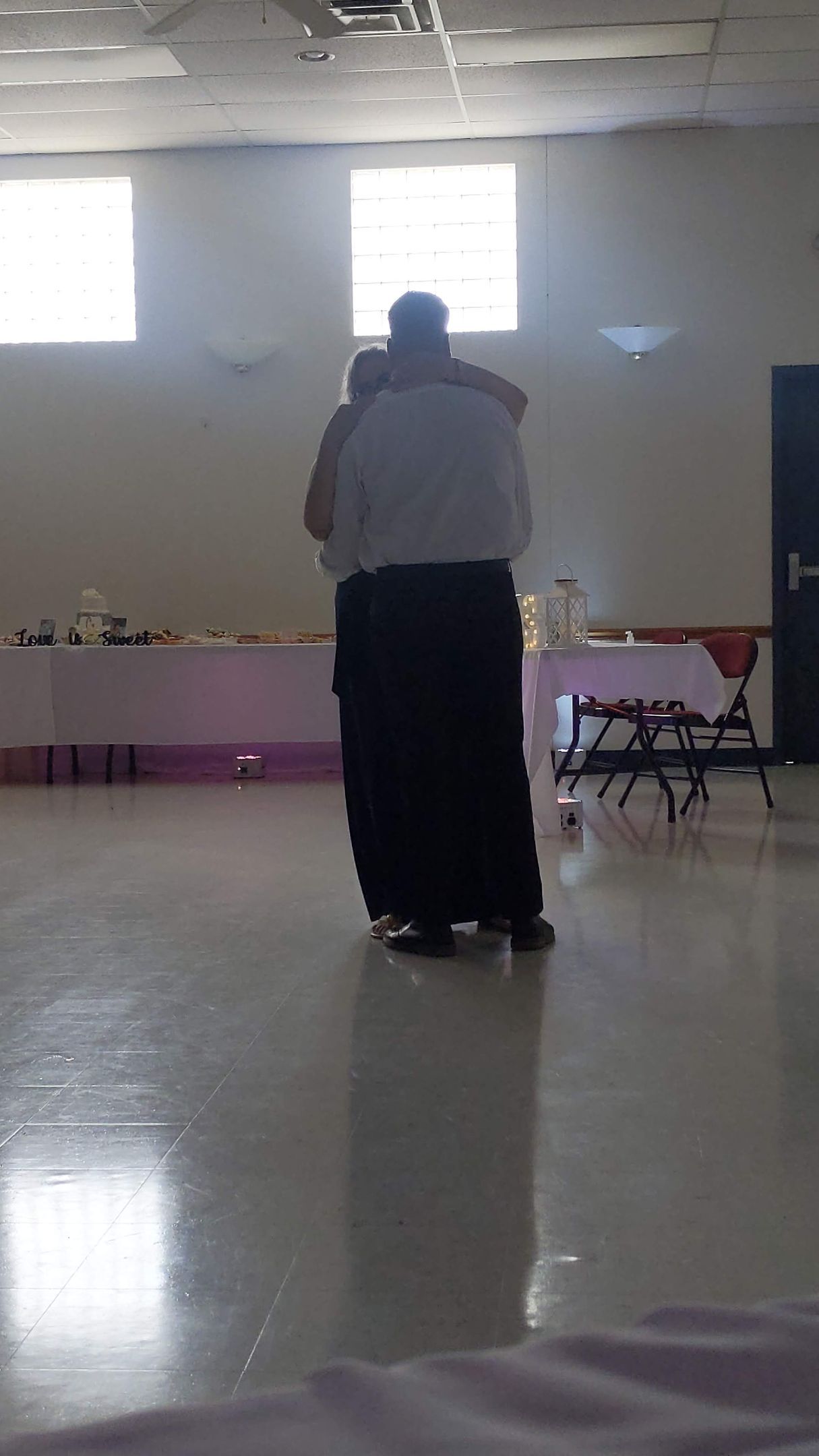 Two people dancing in a bright hall with polished floor and tables along the back wall