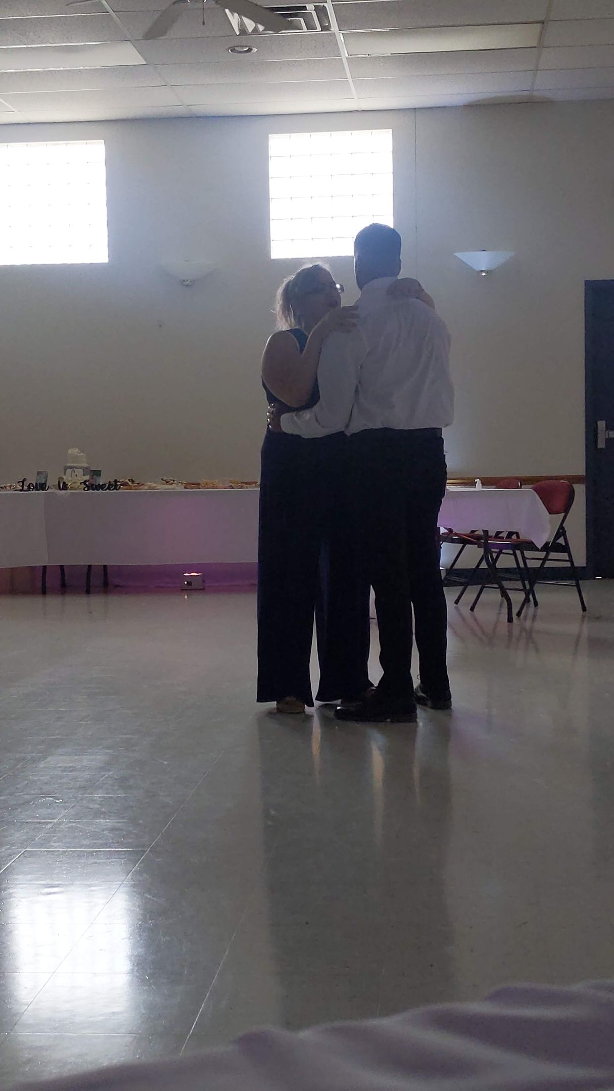 Two people embracing in a bright hall with tables along the back wall.