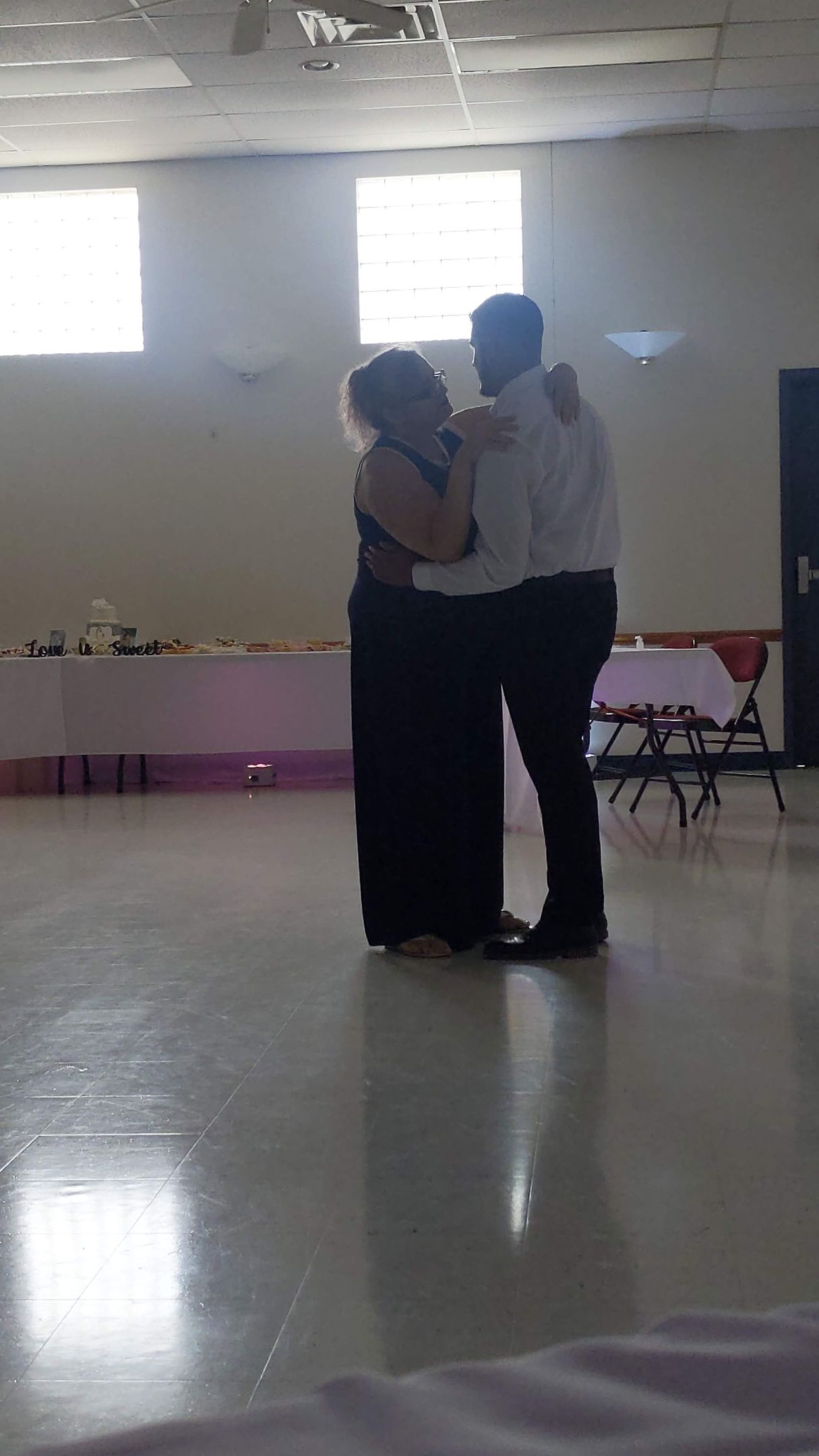 Two people dancing closely in a bright hall with high windows and a polished floor.