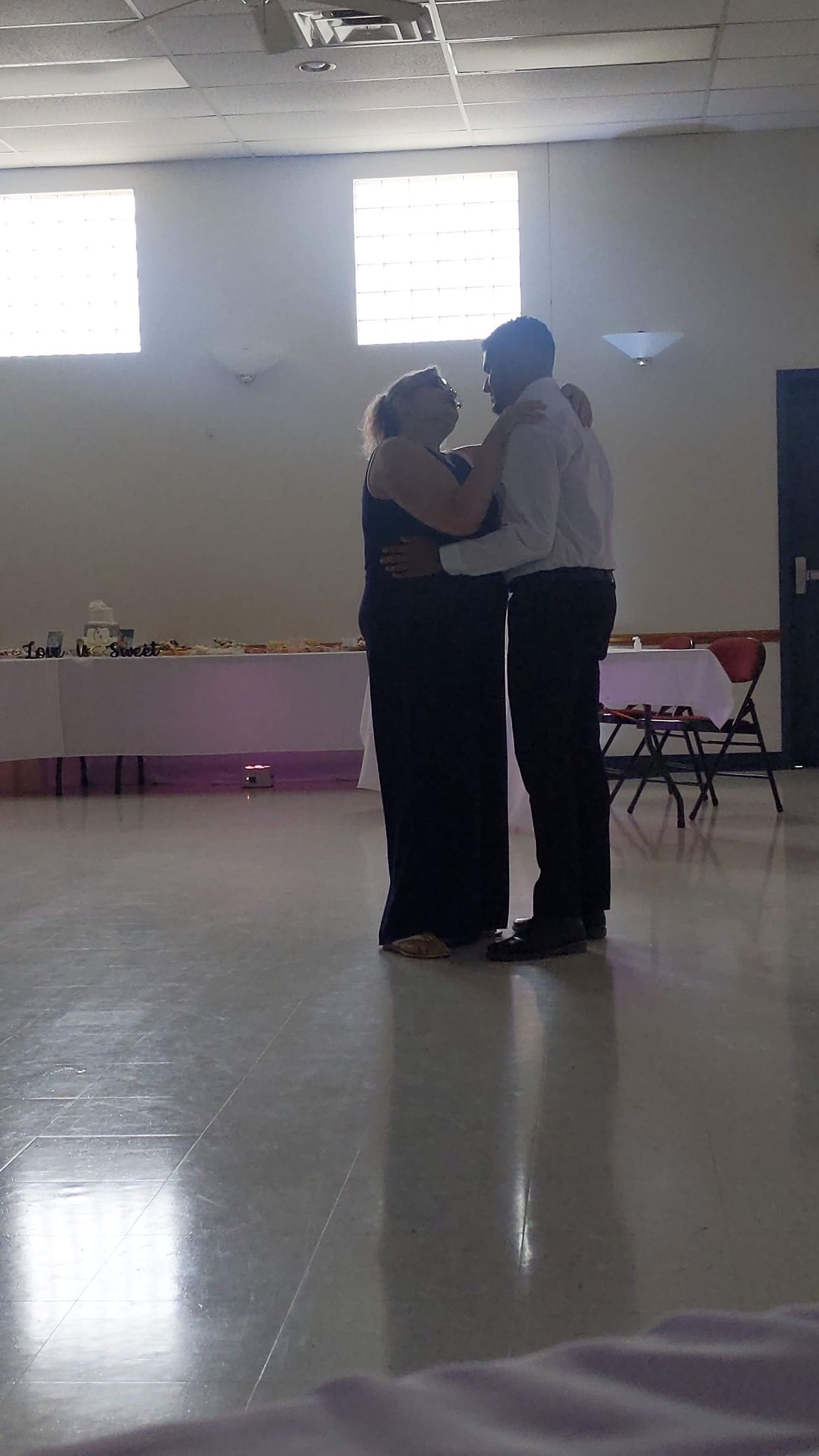 Two people embrace on a polished dance floor in a bright hall with tables along the wall.