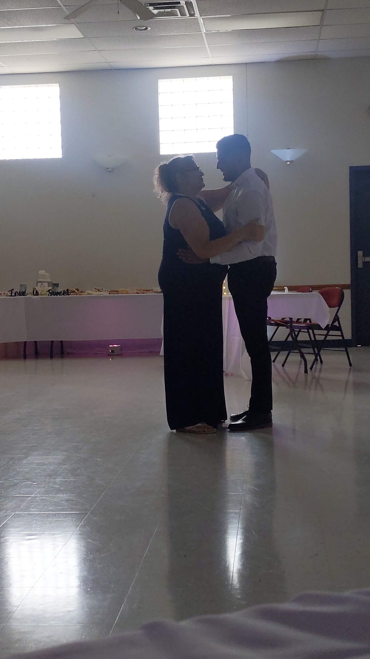 Two people dancing closely in a bright hall with windows and a polished floor.