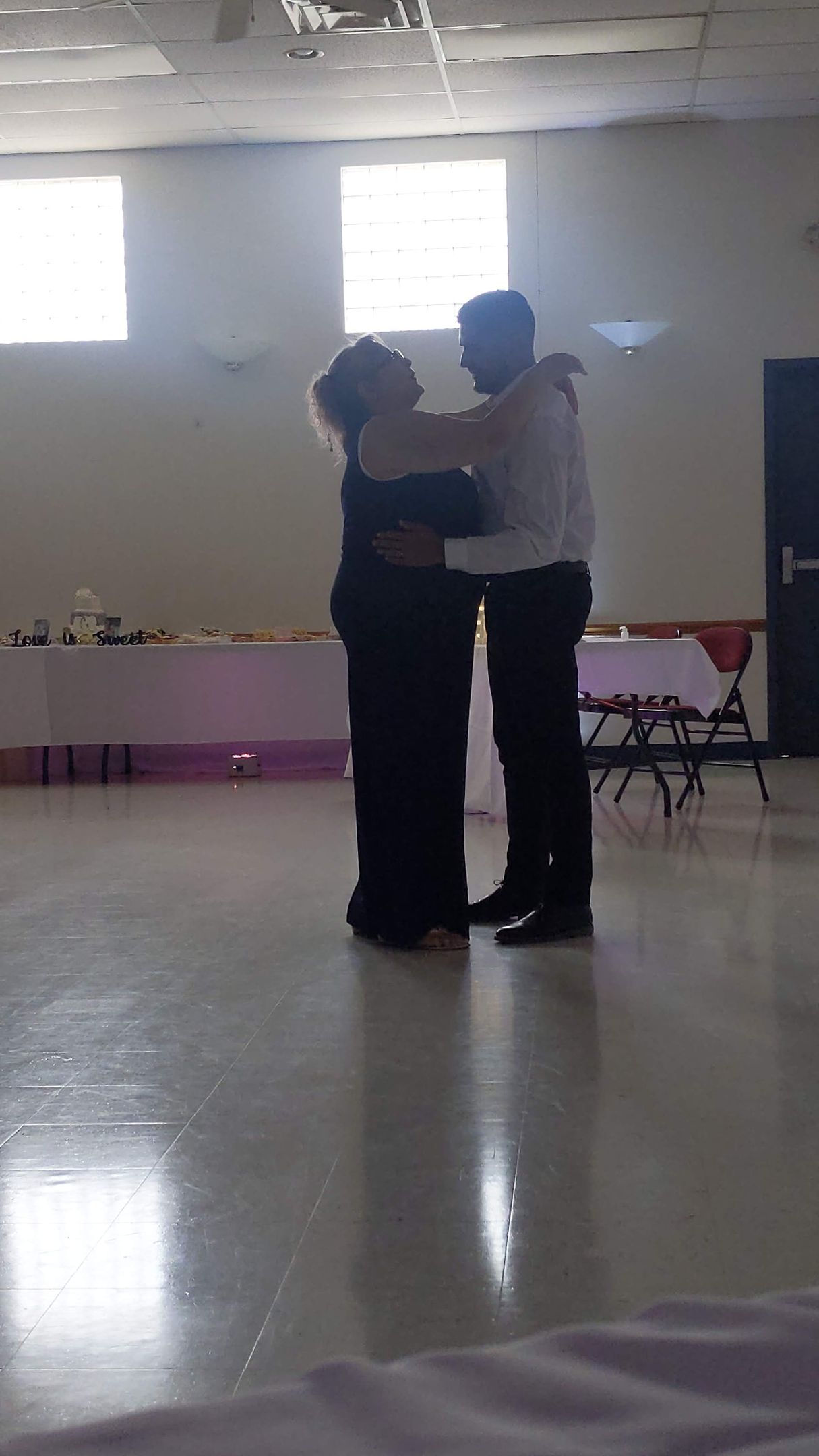 Two people dancing closely in a bright room with windows and a polished floor.