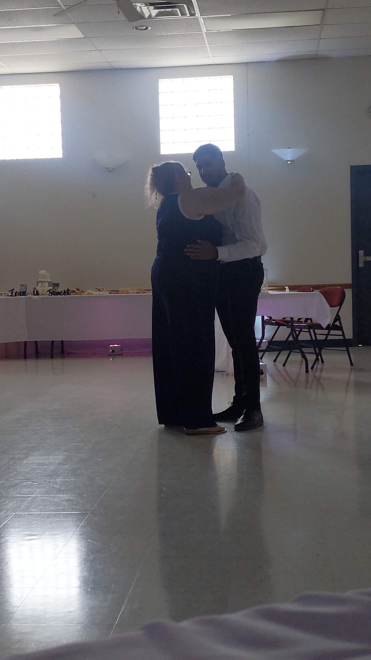 Two people embracing in a bright hall with tiled floor and tables along the wall.