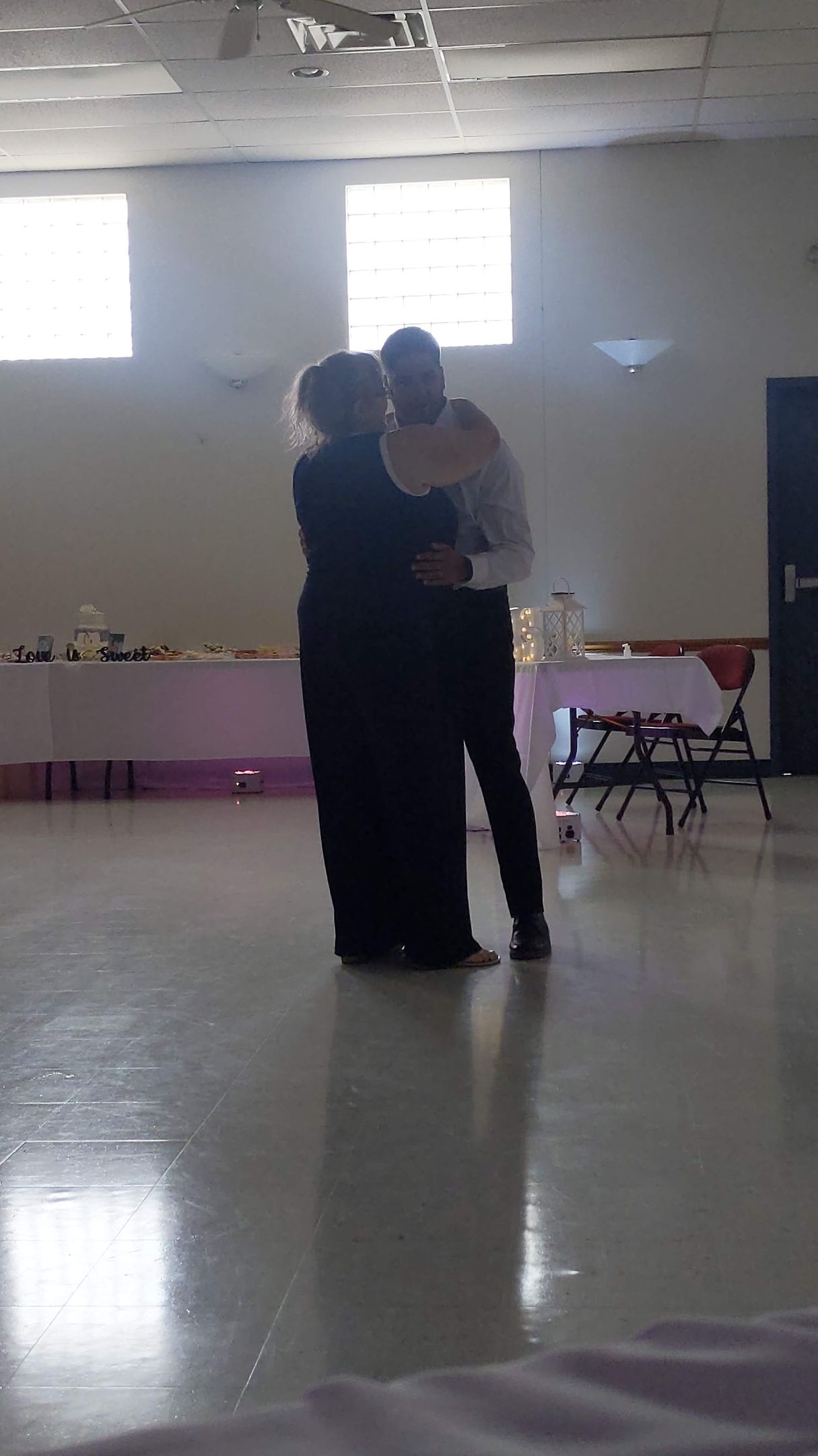 Two people dancing closely in a bright ballroom with polished floors and windows.