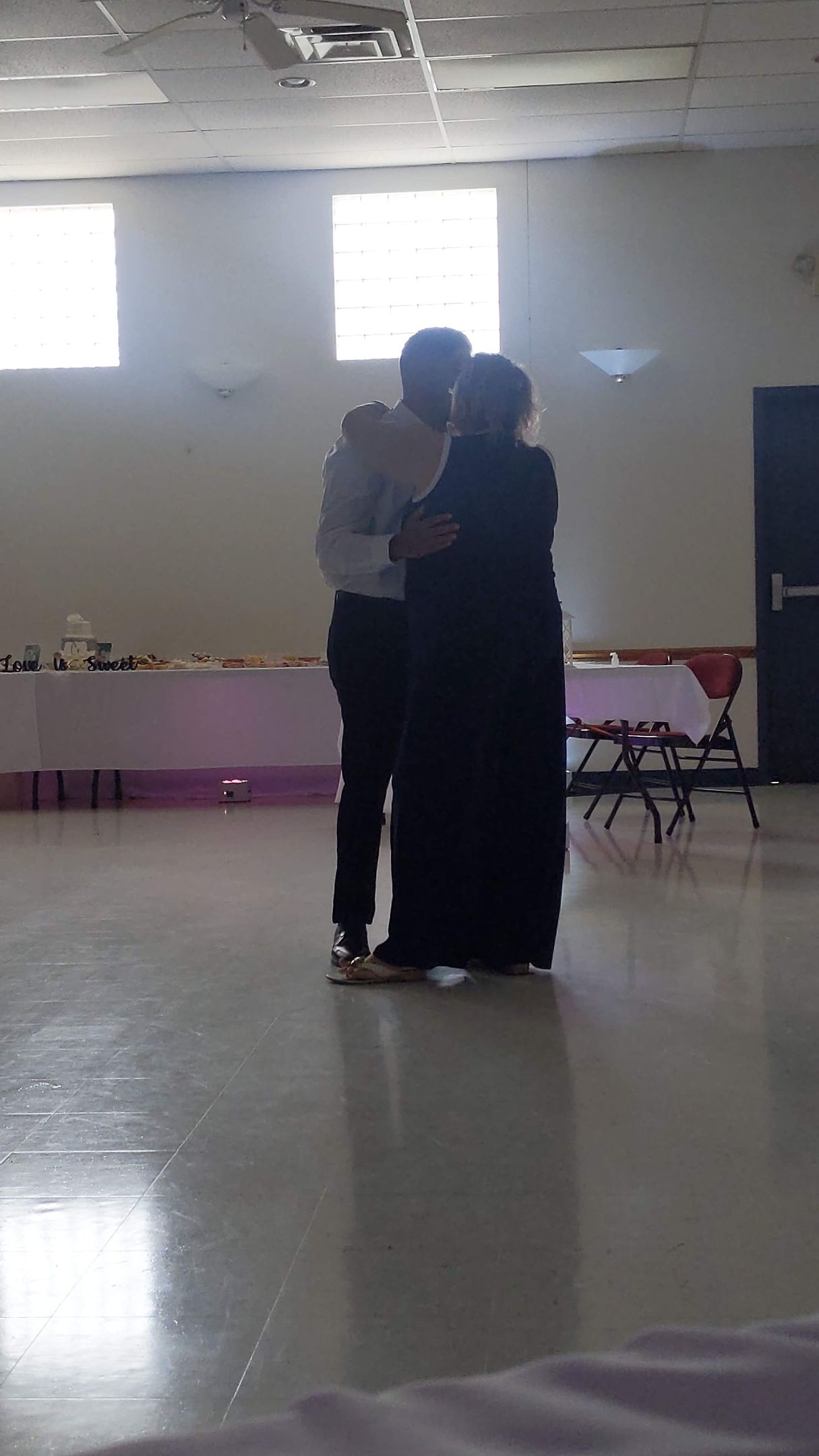Two people embrace in a bright indoor hall with tables along the wall.