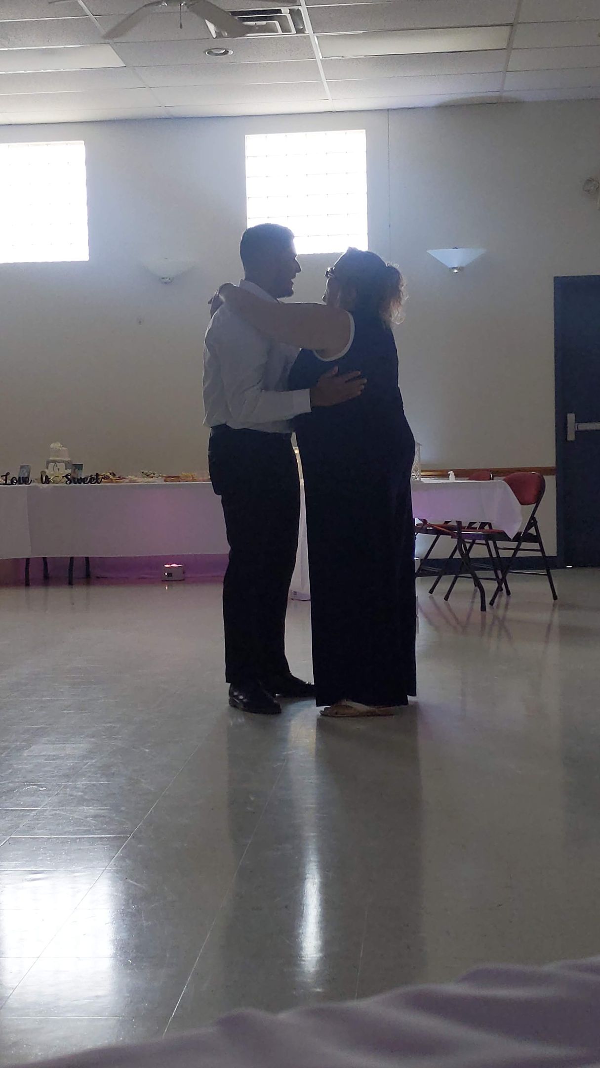 Two people dancing closely in a bright room with a polished floor and tables along the wall.